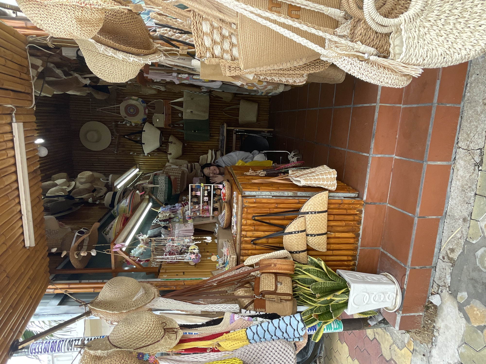 The image depicts the interior of a small shop located in Phu Thuy Ward, Vietnam. The shop is filled with straw hats and woven bags, hanging from the ceiling and displayed on shelves. A person is lying on a surface in the back of the shop, seemingly resting. The shop's structure appears to be primarily made of bamboo and wood. The floor is tiled with square terracotta tiles. A potted plant stands near the entrance.
