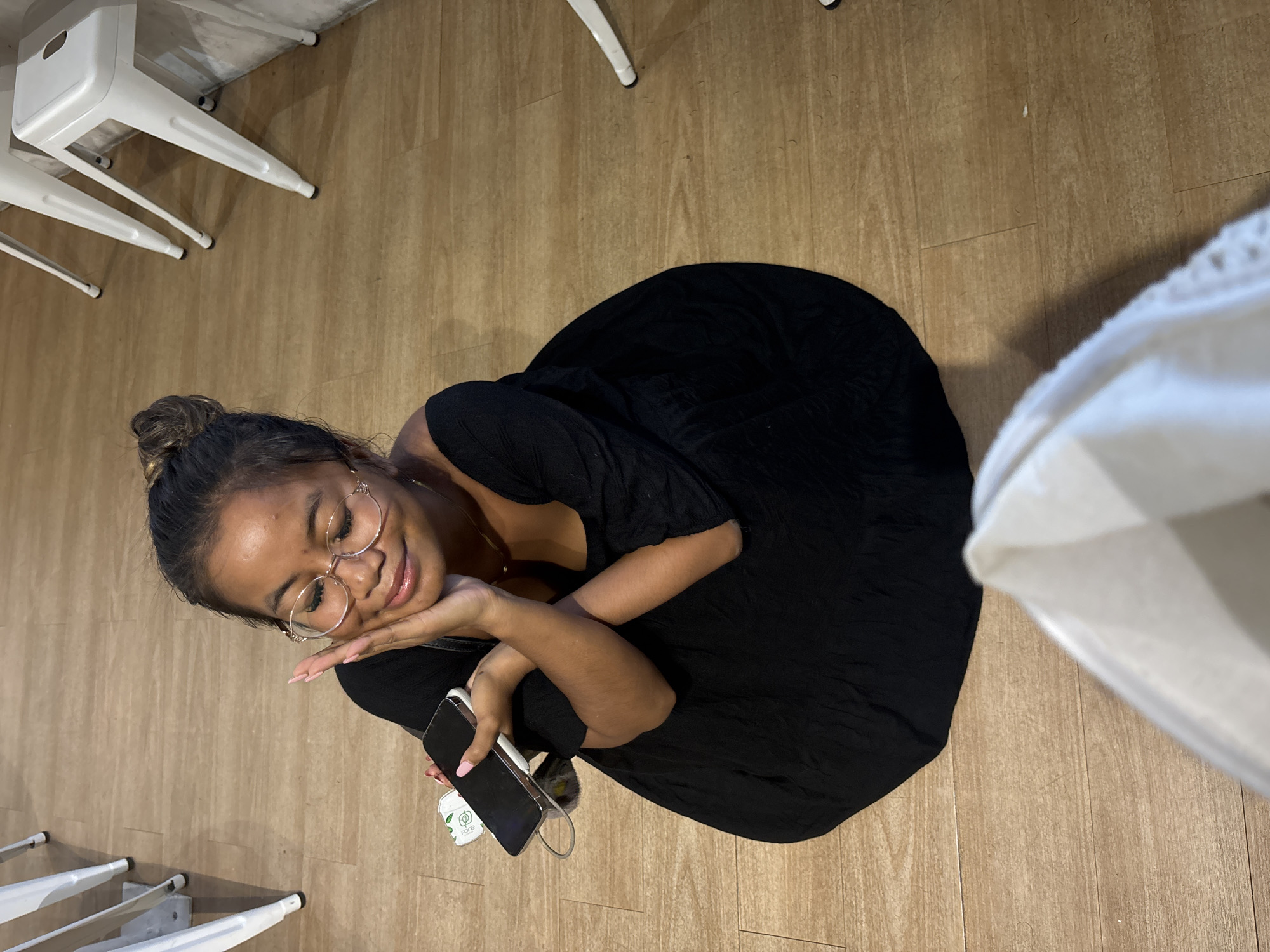 The image shows a woman with her hair in a bun, wearing glasses and a black dress. She is resting her head on her hand and holding a phone. There are white chairs nearby, and the floor appears to be made of wood. Given the presence of wood and white furniture, the location is plausibly an indoor establishment such as a restaurant or a café. Because no geographical details are provided in the image, it is not possible to confirm whether the location is in North Jakarta, Indonesia, or not.