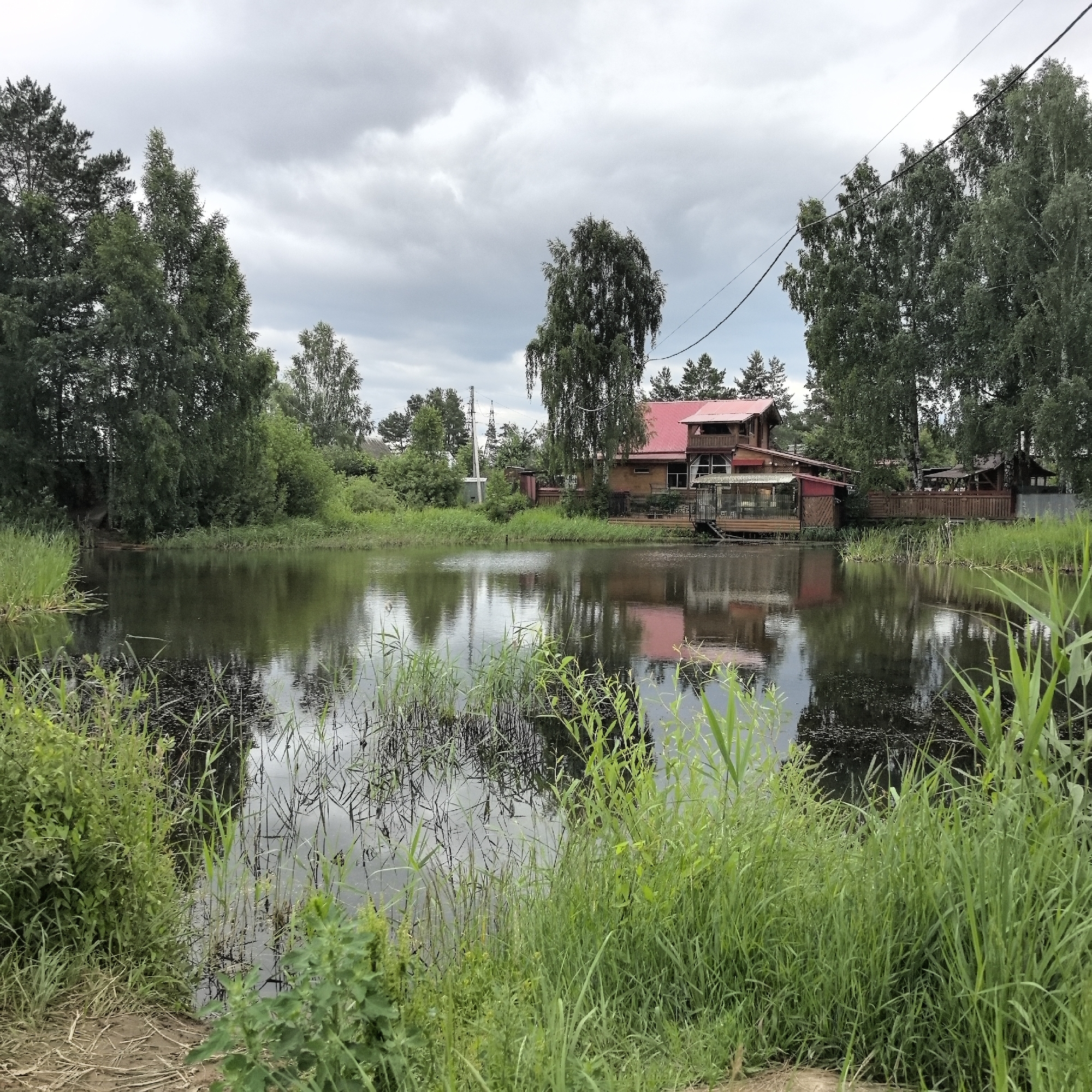 This media file depicts a tranquil, natural scene in Noviy, Russia, under a cloudy sky. The foreground is dominated by lush, green grass and reeds growing along the edge of a dark, reflective pond. The pond's surface mirrors the overcast sky and the surrounding trees, creating a slightly somber yet peaceful atmosphere.
In the mid-ground, a wooden house with a vibrant red roof stands prominently. The house appears to be multi-story and features balconies and large windows, suggesting it might be a residence or a guesthouse. It is nestled amongst a variety of trees, including tall pines and birches, which line the perimeter of the property and the pond. The overall setting is rural and serene, with elements of both natural beauty and human habitation.
The weather appears to be overcast, contributing to the muted lighting of the scene. The clouds are thick and grey, hinting at the possibility of rain, but no precipitation is visible. The time of day is difficult to ascertain precisely due to the diffused light, but it seems to be daytime. There are no people or discernible activities taking place; the image captures a still moment in time, emphasizing the quietude of the location.
Notable details include the reflections on the water, the varied textures of the foliage, and the architectural style of the house. There is no visible text within the image. The scene evokes a sense of calm and natural beauty, characteristic of a rural landscape in Russia.