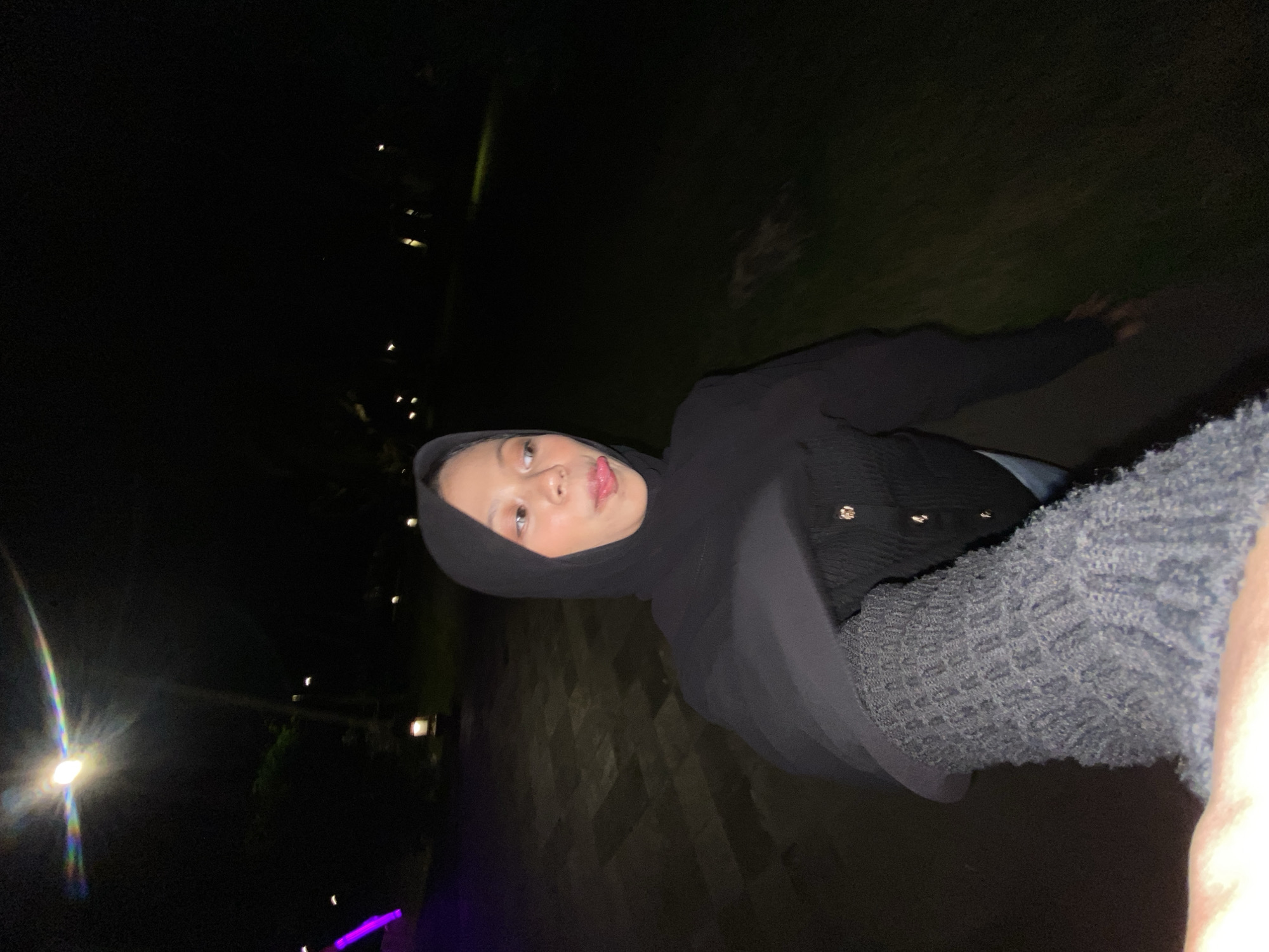 This media file shows a young woman in a black hijab and cardigan posing for a selfie at night. She is sticking her tongue out playfully, and her eyes are wide open. The background is dark, with some blurry lights visible in the distance, suggesting an outdoor setting like a park or a street. There is a cobblestone path in the foreground and some greenery behind the woman. The woman's clothing appears to be a black knitted cardigan over a black hijab. She is wearing a gray, textured scarf or wrap over her shoulder. The overall mood of the image is casual and lighthearted, with the woman appearing to be having fun. The time of day is clearly night, and there are no indications of specific weather conditions. There is no visible text in the image. The location context is Bulusan, Indonesia.