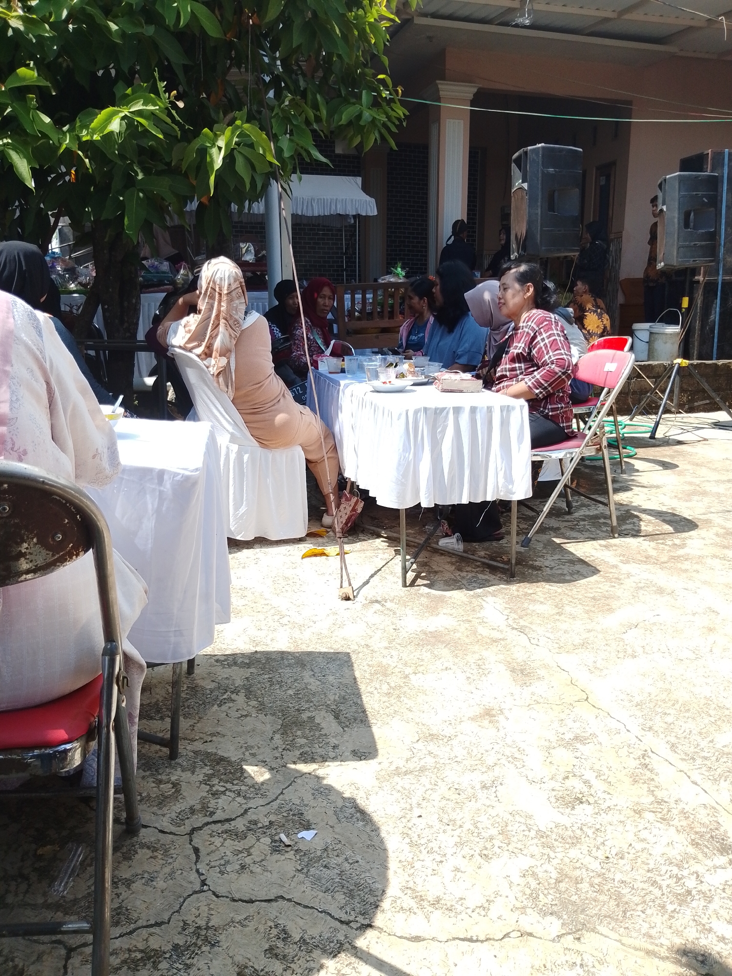 This image shows an outdoor scene with a gathering of people in Gabus, Indonesia. Several tables covered with white tablecloths are set up, and people are seated around them, possibly eating or socializing. Most of the individuals are women, with some wearing hijabs. There are additional people standing or walking in the background. A tree provides shade in one area. In the background, there appears to be a building, and speakers are visible. The ground is a cracked concrete surface. There is litter in the lower right. The overall atmosphere suggests a community event or celebration.