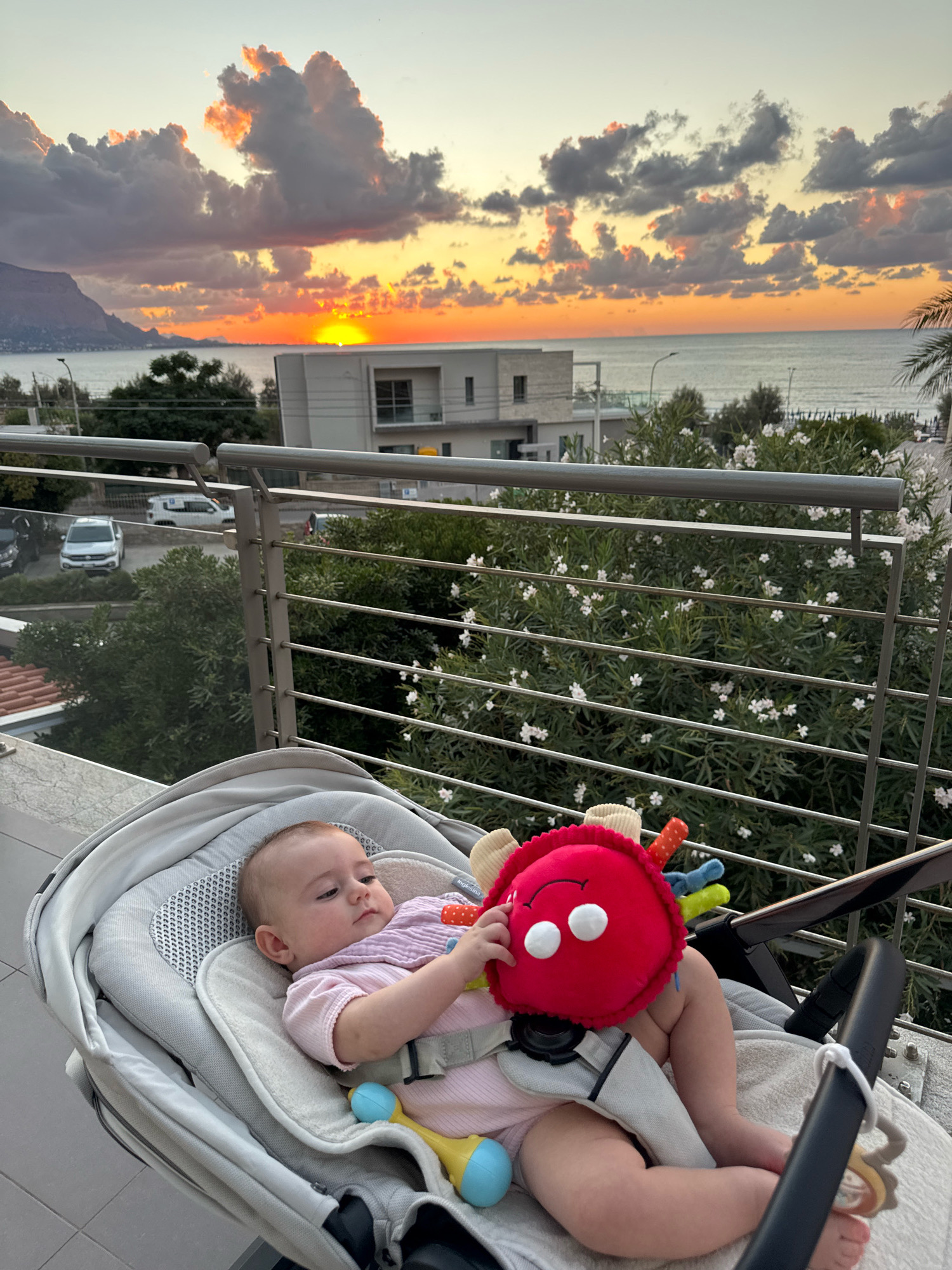 A baby in a stroller plays with a red toy on a balcony overlooking the sea at sunset in Capaci, Italy. The baby is wearing a pink outfit and is propped up in a gray stroller. The toy is red and plush with white eyes and a smiling mouth. In the background, the sun is setting over the Mediterranean Sea, casting a warm glow on the clouds. Buildings and trees are visible along the coastline. The balcony railing is made of metal.