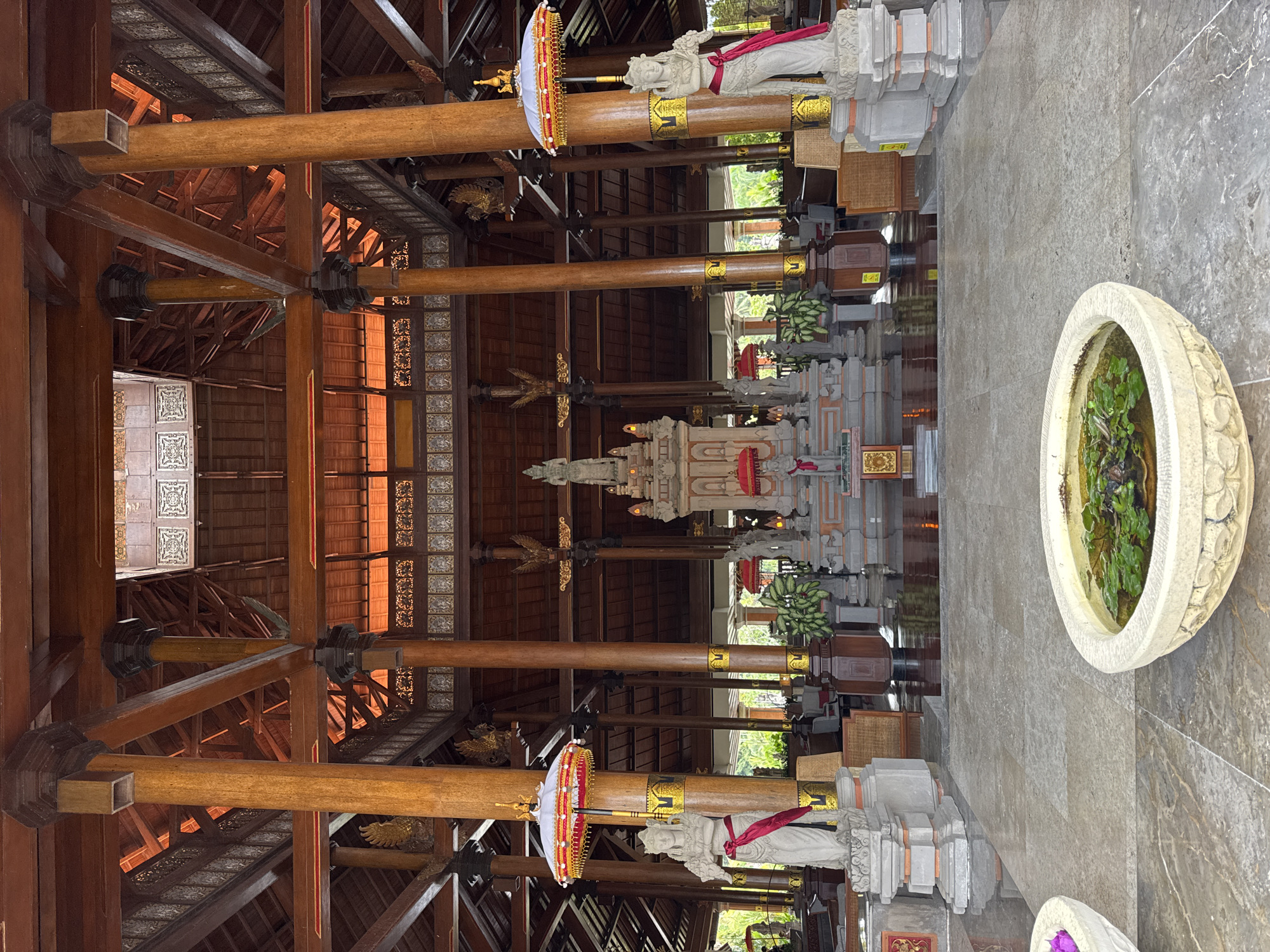 The image captures the interior of a grand hall, likely a lobby or reception area, in Kuta Selatan, Indonesia. The architecture is characterized by rich, dark wood used for the extensive ceiling structure, supported by numerous tall, circular wooden pillars. Intricate carvings and decorative elements are visible throughout the wooden beams and ceiling panels.
Dominating the center of the hall is a multi-tiered, ornate altar or shrine, featuring stone statues and red umbrellas. Flanking the altar are two large stone statues of women, each adorned with a vibrant red sash and holding what appears to be flowers. Large, decorative umbrellas, similar to the ones near the altar, are positioned on ornate stands next to these statues.
The floor is paved with large, polished stone tiles, reflecting the warm light filtering from above. In the foreground, a shallow, decorative stone basin filled with water and aquatic plants sits on the floor. In the background, glimpses of lush greenery and outdoor elements are visible through open doorways or windows, suggesting a tropical setting. The overall ambiance is one of grandeur, cultural richness, and tropical serenity.