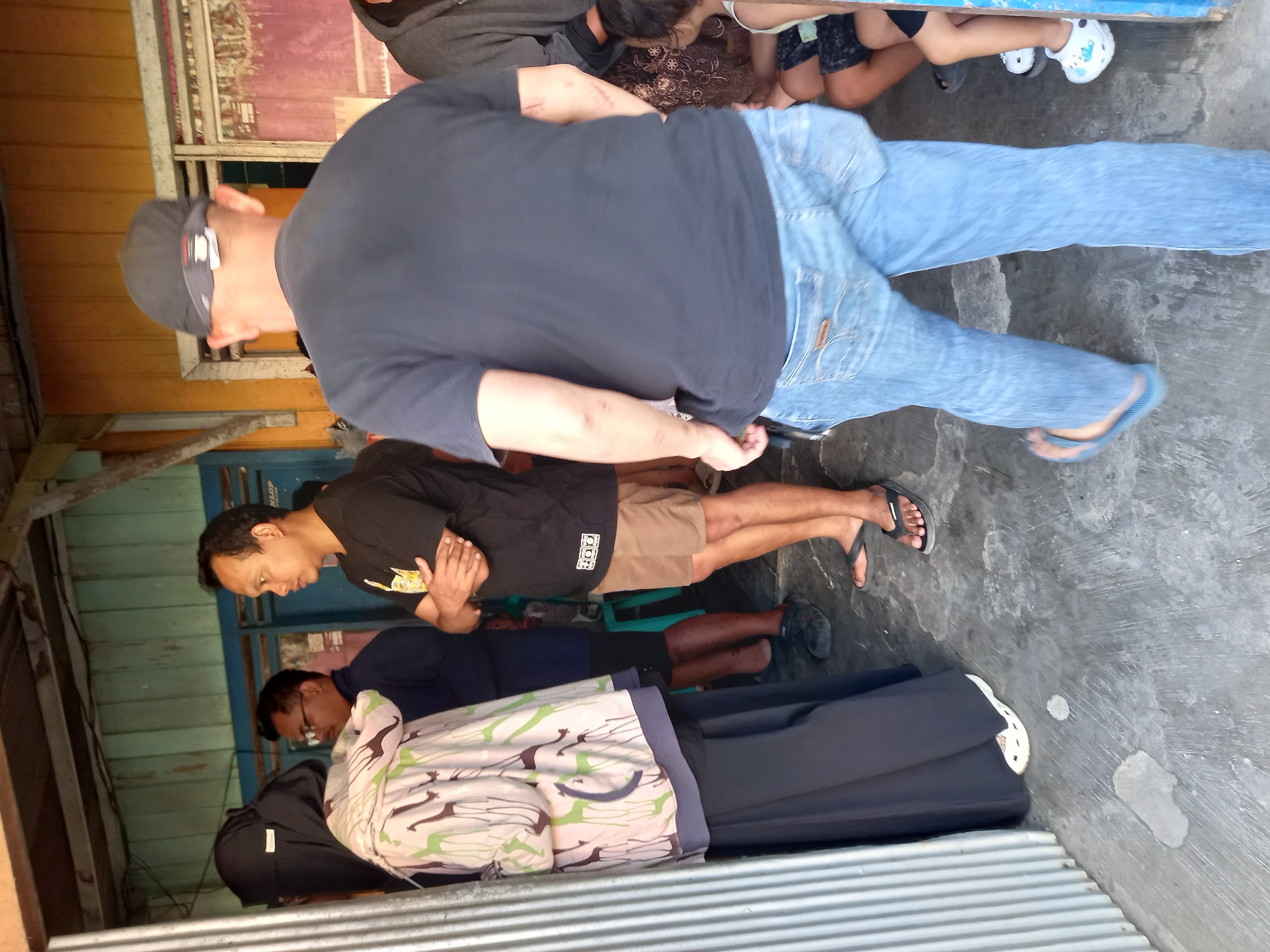 The image captures a street scene in Samarinda, Indonesia, depicting a group of people gathered near a building entrance. A woman wearing a black hijab and a patterned cardigan stands to the left. Beside her, a man in a navy blue shirt and glasses looks away. In the foreground, a man in a black t-shirt and blue jeans walks away from the camera. To the right, a child in white Crocs is visible. The environment appears to be an outdoor area with a weathered building featuring wooden and metal elements. The ground is concrete, with some debris and water stains. The lighting suggests it is daytime.