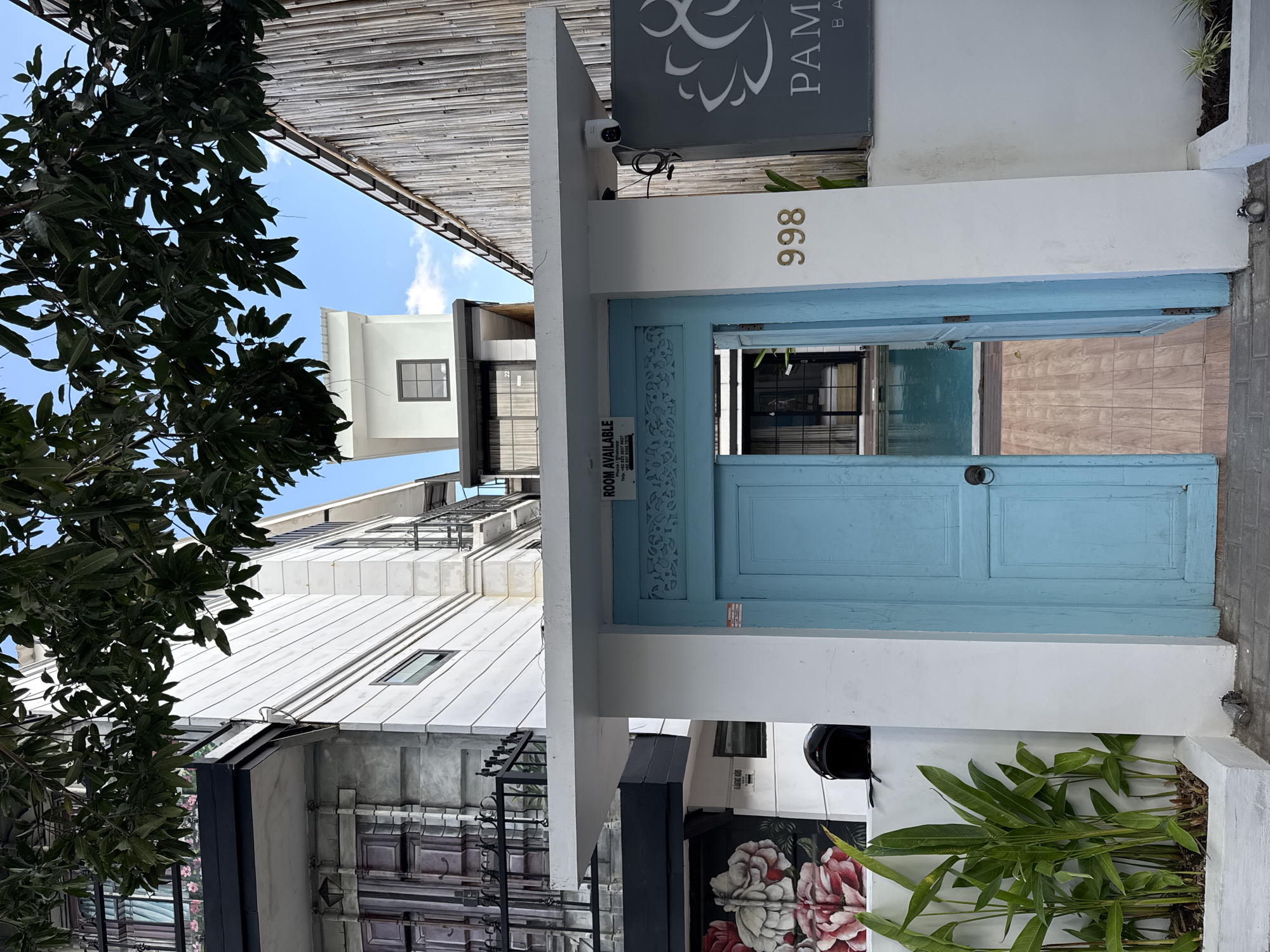The image depicts the entrance to a building, likely a guesthouse or hotel, located in the city of North Kuta, Indonesia. A bright turquoise door, partially ajar, reveals a glimpse of a swimming pool and a wooden pathway. Above the door, a white awning extends, supporting a sign that reads "ROOM AVAILABLE" and providing contact information. A security camera is mounted on the underside of the awning. To the right of the door, the number "998" is affixed to a white pillar, adjacent to a dark sign bearing a logo and the text "PAME BA". The surrounding buildings are modern with white facades, featuring balconies and windows. Lush green plants are visible in planters near the entrance, and the scene is framed by the leaves of a large tree overhead. The sky is clear and blue.