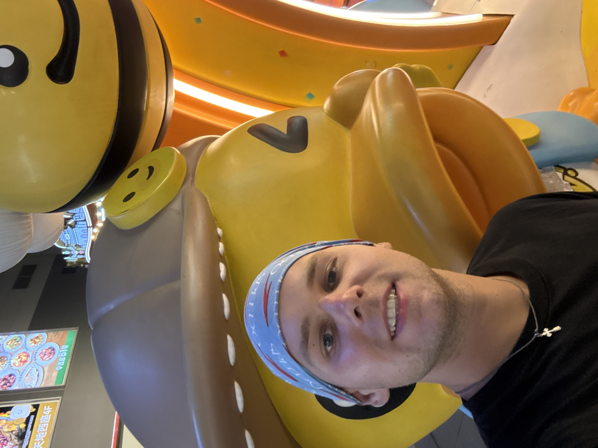 A young Caucasian male wearing a black t-shirt and a blue bandana stands in front of a large yellow cartoon duck statue. The duck statue is wearing a brown hat and has a large beak. Behind the duck, a large yellow bee statue is partially visible. The background appears to be an indoor shopping mall with neon lights and advertisements. The scene is likely in the city of Tianhe District, China, based on the advertisements in Chinese text visible in the background. The male is smiling and looking at the camera, with a silver cross necklace around his neck.