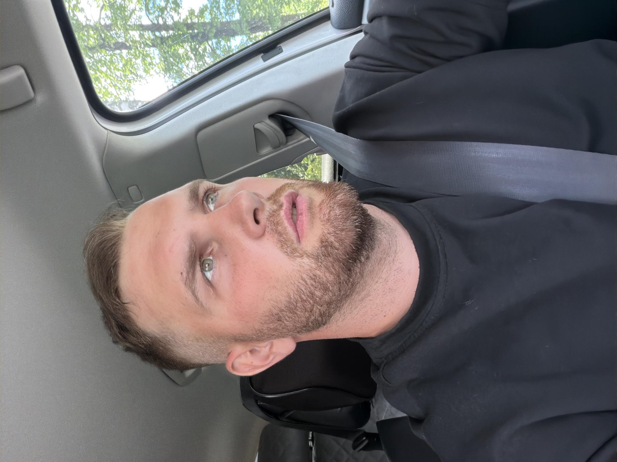 The image presents a close-up, slightly rotated view of a light-skinned man inside a vehicle, likely in Chaikovsky, Russia. The man has short light brown hair, a short beard, and striking green eyes. He is wearing a black long-sleeved shirt and a dark grey seatbelt that crosses his chest. His head is reclined against a black headrest, and he is looking upwards with a neutral, slightly contemplative expression, his mouth slightly ajar.
The scene is set within a car, featuring a light grey interior headliner and paneling. Above the man, a sunroof or large window reveals a bright blue sky and lush green tree foliage, indicating it is daytime with sunny weather. The natural light illuminates the man's face and the car's interior. The visible elements suggest the man is either resting or observing the surroundings while in transit or parked. No text is visible in the image.