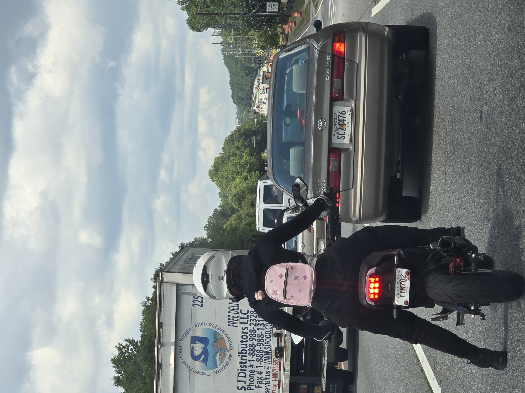 A man wearing a white helmet, black jacket, and black pants rides a black motorcycle. A person wearing a black hooded sweatshirt and black pants is seated behind the man on the motorcycle. The passenger is wearing a small pink backpack with a floral pattern. The motorcycle's taillight is illuminated red. The license plate on the motorcycle reads "GEORGIA YYM702".
In front of the motorcycle is a tan Toyota Camry with a Georgia license plate that reads "SCZ4476". The Toyota Camry has its brake lights illuminated. To the left of the motorcycle is a white semi-truck with "SJ Distributors LLC" printed on its side.
The vehicles are on a multi-lane paved road with white lane markings. The environment appears to be a highway or a major road in Alpharetta, Georgia, United States of America, with trees and power lines visible on the sides of the road. The sky is cloudy with patches of blue.