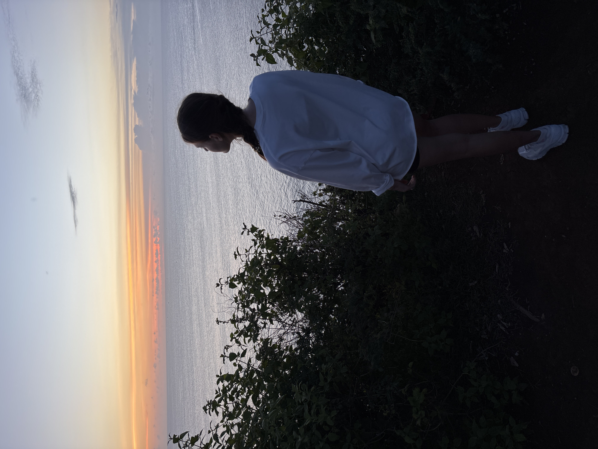 A young woman with a braid stands on a cliff overlooking the ocean at sunset. The sky shows a gradation of colors from light blue to orange. The location is Pecatu, Indonesia, identifiable by the geography and coastal landscape. Bushes line the edge of the cliff. The woman is wearing a white long-sleeved shirt, shorts, and white sneakers. She is facing the ocean.