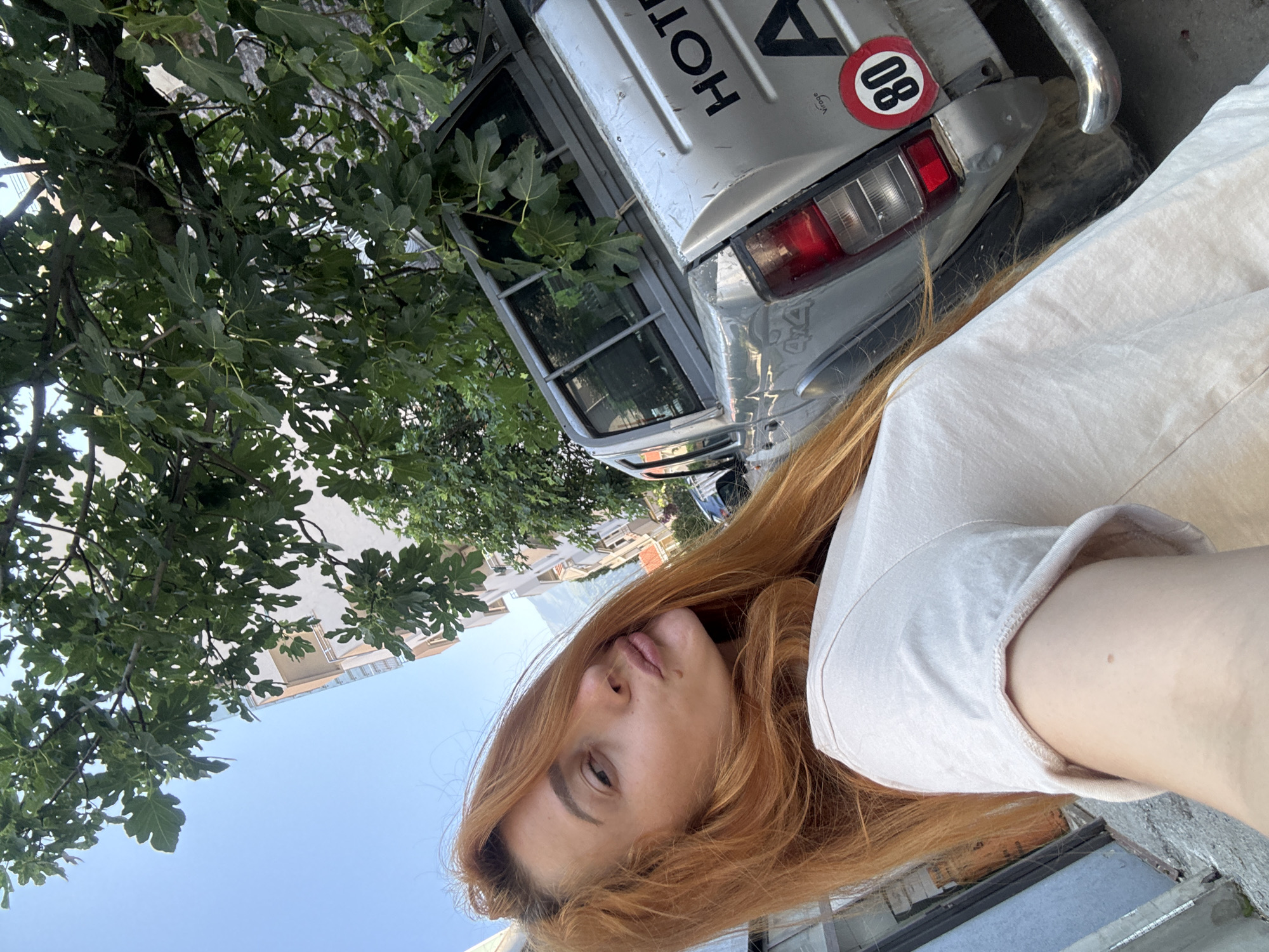 A woman with auburn hair is taking a selfie outdoors. She is wearing a light-colored t-shirt and has her lips pursed. Behind her, a silver pickup truck is parked. The truck has "HOTEL" and "A" visible on its tailgate, along with a circular sticker with "80" in it. A large tree with green leaves overhangs the scene. In the background, a building and a clear blue sky are visible. The scene appears to be in the city of Budva, Montenegro.