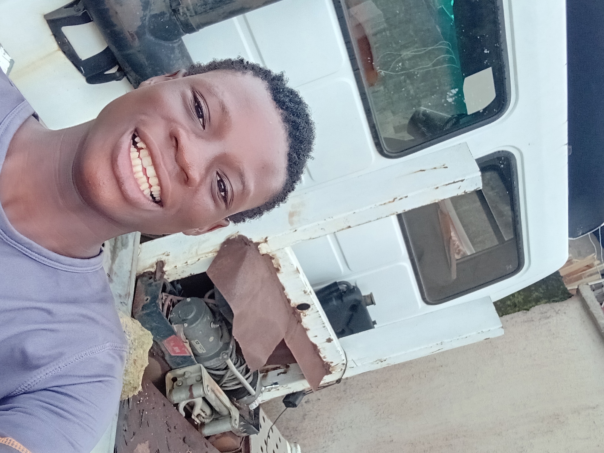 A young male is taking a selfie in front of a white truck. He is smiling and wearing a grey t-shirt. The truck appears to be old and rusty, with some of its mechanical parts exposed. The background shows a textured wall and some greenery. The image is taken outdoors, possibly in an urban or industrial setting, given the presence of the truck and the wall. The lighting suggests it is daytime.
