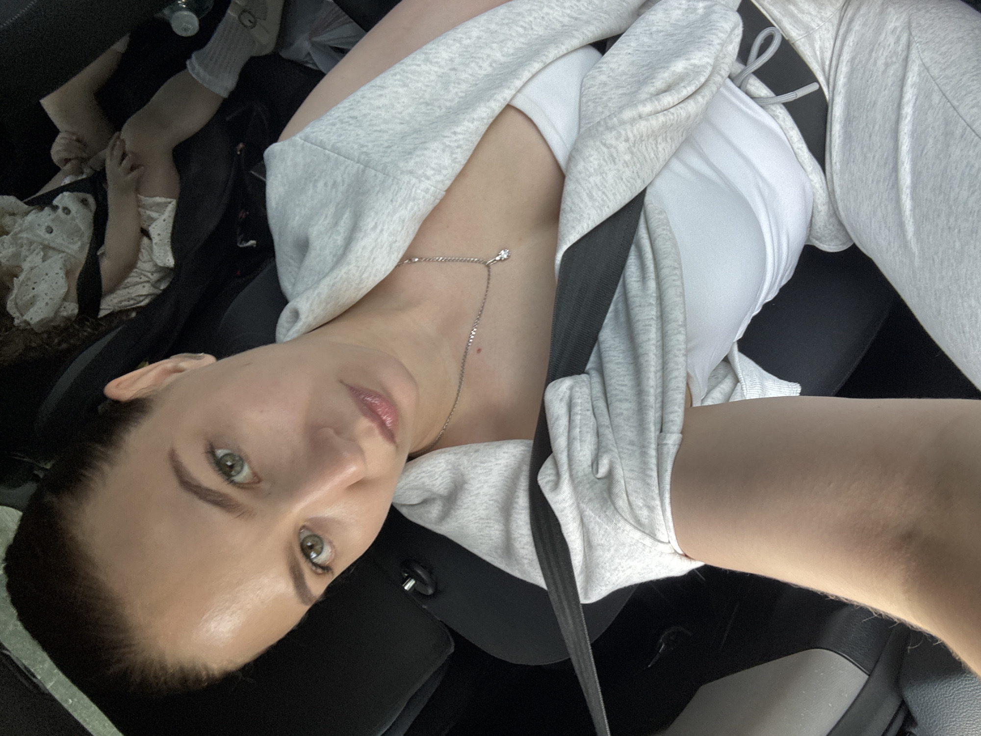 A selfie shot in a car, interior is visible with dark upholstery and seat belt across the person. A person in the driver seat looking at the camera, wearing a white top and grey hooded jacket, and a delicate silver necklace. In the back seat, there is a child with a white frilly dress and white socks. The location is Chaikovsky, Russia.