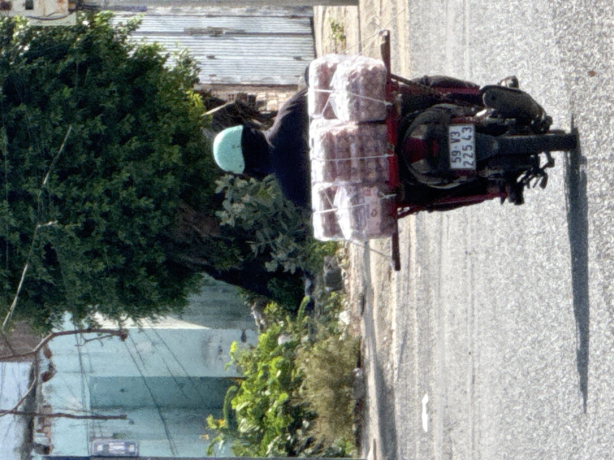 A person wearing a light blue helmet and dark clothing is riding a red motorcycle on an asphalt road. The motorcycle is carrying a large load of white packages, secured with white cord, on its rear rack. The license plate on the back of the motorcycle reads "59-V3 225.43". To the left of the road, there is a light blue building with a rough texture, and further back, a large, dark green tree with dense foliage. To the right of the road, there is a corrugated metal fence. The lighting suggests it is daytime.