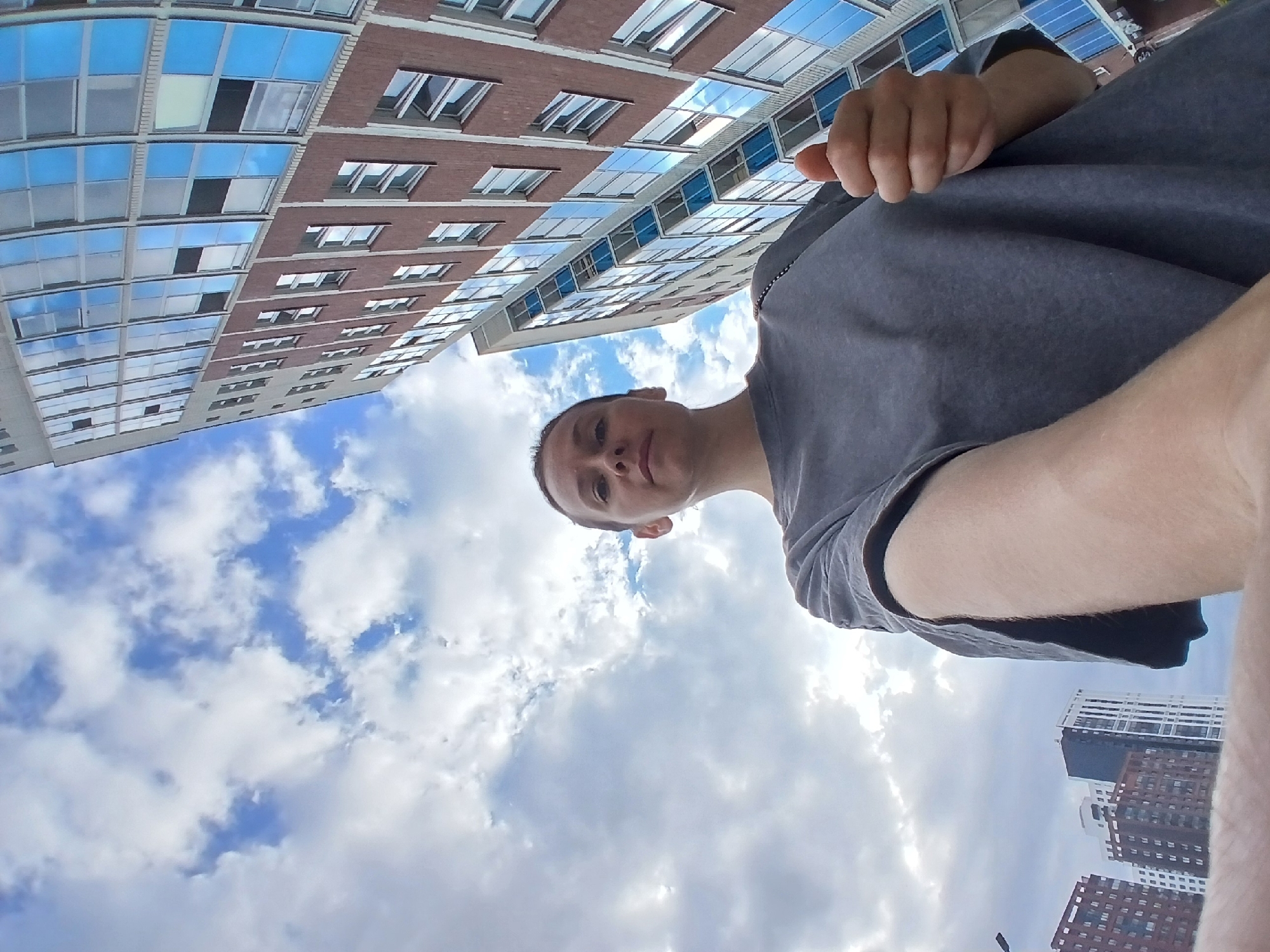 In Yekaterinburg, Russia, a low-angle shot captures a woman standing outdoors with a wide-angle lens, emphasizing the sky and surrounding buildings. She is positioned in the foreground, looking towards the camera with a neutral expression. Her short, dark hair is pulled back, and she is wearing a grey t-shirt. Her right arm is extended, and she is giving a thumbs-up gesture. Behind her, two modern, multi-story brick and glass buildings rise up, their reflective windows mirroring the cloudy sky. In the distance, more apartment buildings are visible. The overall scene suggests a casual, urban environment under a partly cloudy sky.