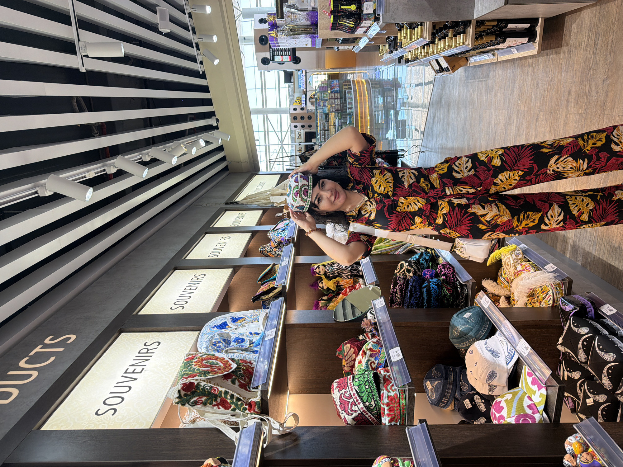 A woman, with dark hair and wearing a colorful floral jumpsuit and a matching hat, stands in a souvenir shop in Samarkand, Uzbekistan. She is posing for a photo, holding a traditional Central Asian hat on her head. The shop displays a variety of souvenirs, including embroidered hats, bags, and dolls. The shelves are well-lit with overhead spotlights, and the store appears to be spacious and modern. In the background, other shops and display cases are visible, suggesting a commercial area. The overall atmosphere is bright and inviting, with a focus on cultural merchandise.