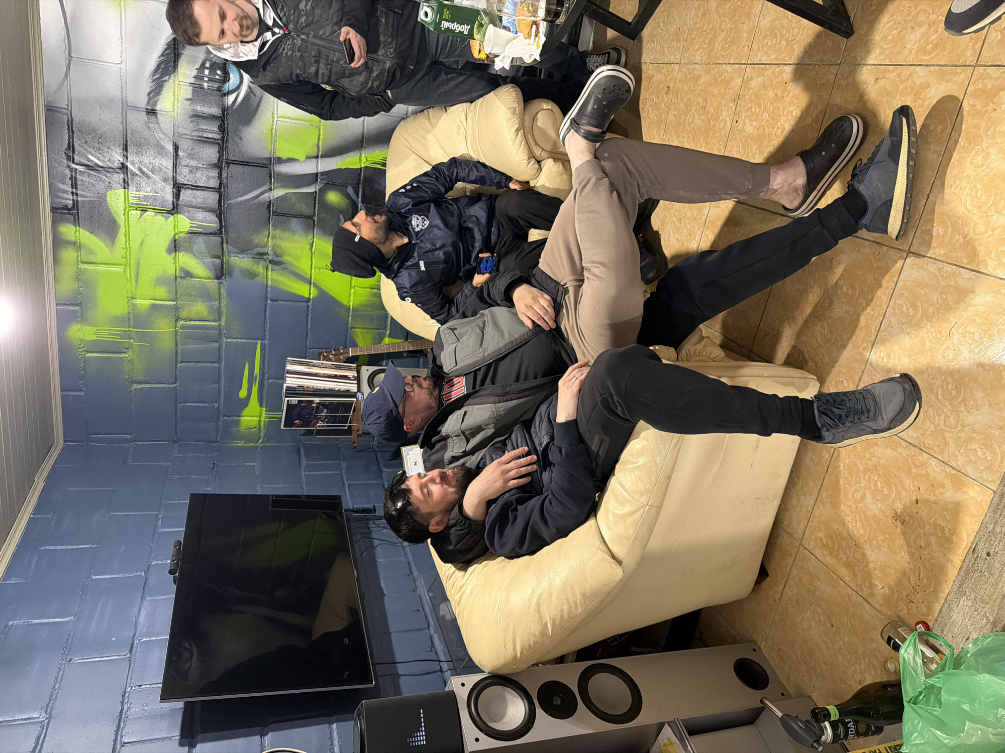 The image depicts an indoor scene, possibly in Ust-Ilimsk, Russia, given the product labels visible. Five men are present. One man stands by a wall with a graffiti mural, using a cell phone. Another man is slumped in a cream-colored chair with his eyes closed. Two other men are in the same chair, one resting on the other, with one man wearing a baseball cap. A fifth man is partially visible, seated in the chair. Various objects are scattered around the floor: empty bottles, a green plastic bag, and speakers. A television is mounted on the wall. A small table is nearby. A guitar is leaning against the wall. The flooring is tiled.