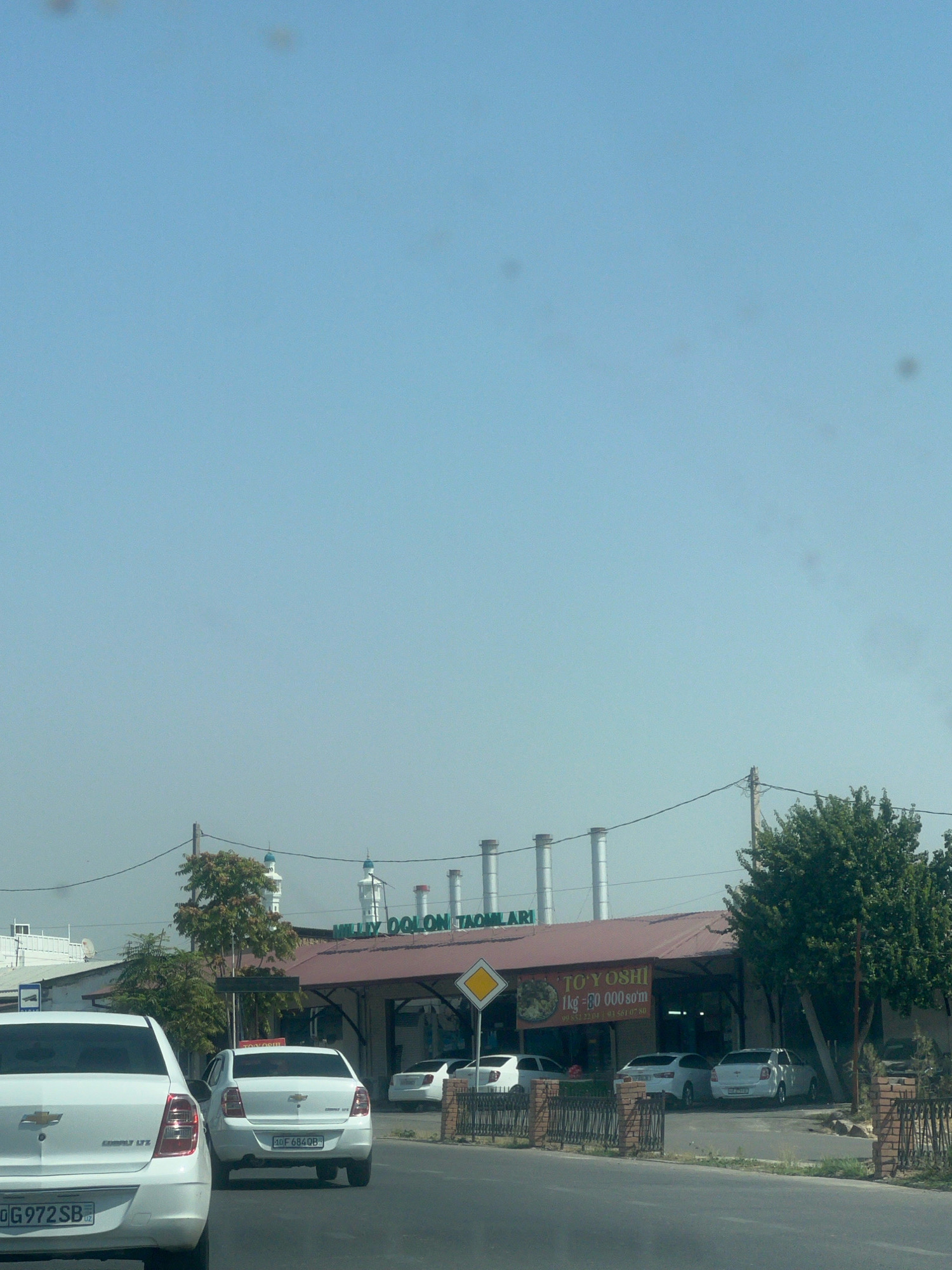 In a street scene in Unknown, Unknown, the foreground features the rear of a white Chevrolet sedan. The car displays the license plate "0G 972 SB". To its right and slightly ahead is another white Chevrolet sedan, this one showing the license plate "10F 684QB". In the middle ground, a low-rise building with a maroon roof is visible, featuring the text "TO'Y OSHI" above an open storefront. To the left of this building, a white tower with decorative elements rises, and further left are more buildings. Several industrial chimneys are present behind the maroon-roofed building. Trees and foliage are scattered throughout the scene, with a particularly large tree to the right of the building. The sky is a clear, light blue. The overall impression is of a commercial area on a bright, clear day.