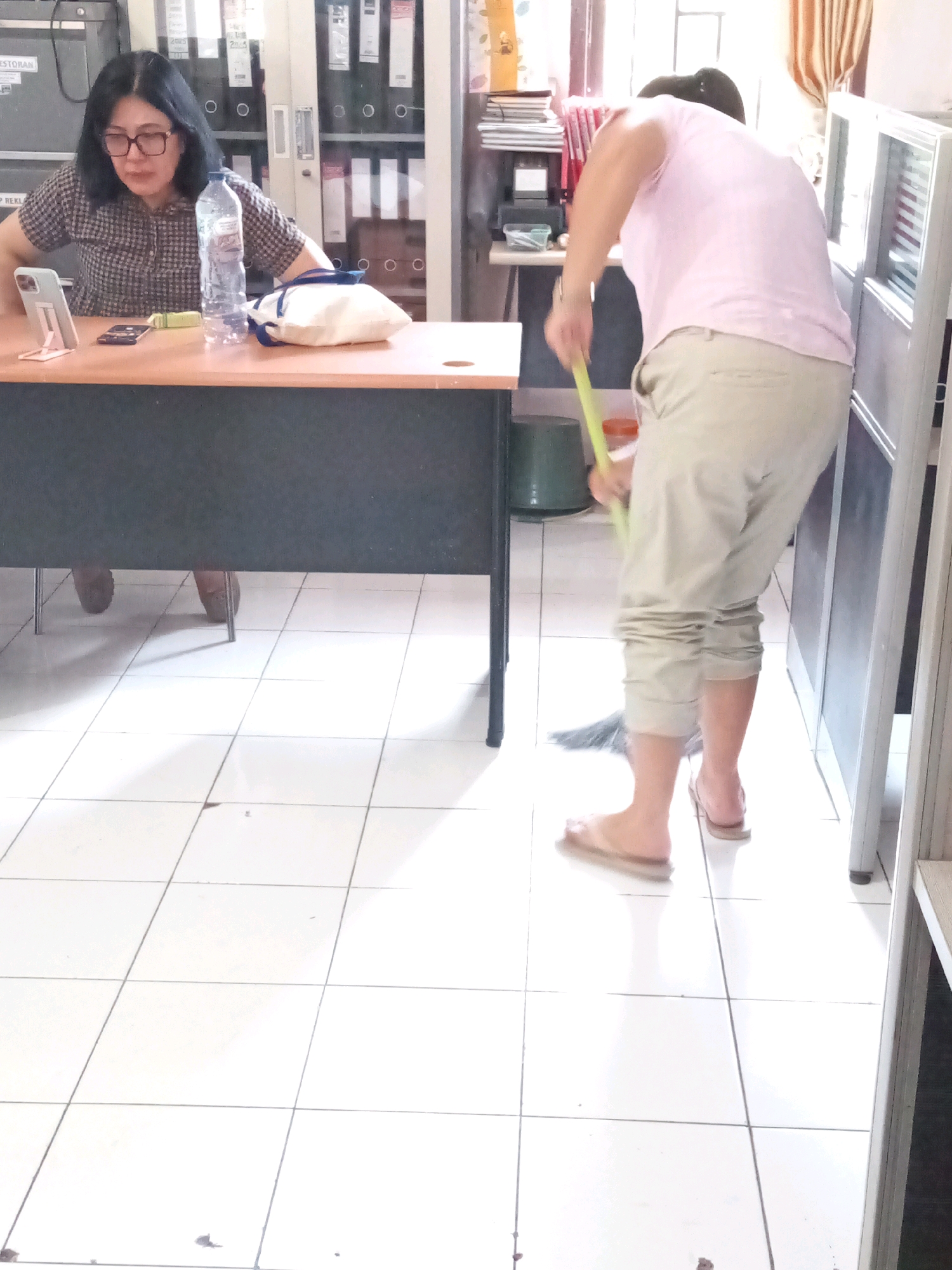 In a brightly lit room with white tiled flooring, a woman with dark hair and glasses is seated at a desk. She is wearing a checkered shirt and appears to be looking at a phone propped up on a stand. A water bottle and a small white bag are also on the desk. To the right of the frame, another person is sweeping the floor with a yellow broom. This individual is wearing a light pink t-shirt and light-colored pants, and is barefoot in simple flip-flops. The background suggests an office environment with filing cabinets and shelving units visible. The overall scene is mundane and domestic, depicting a moment of everyday activity.