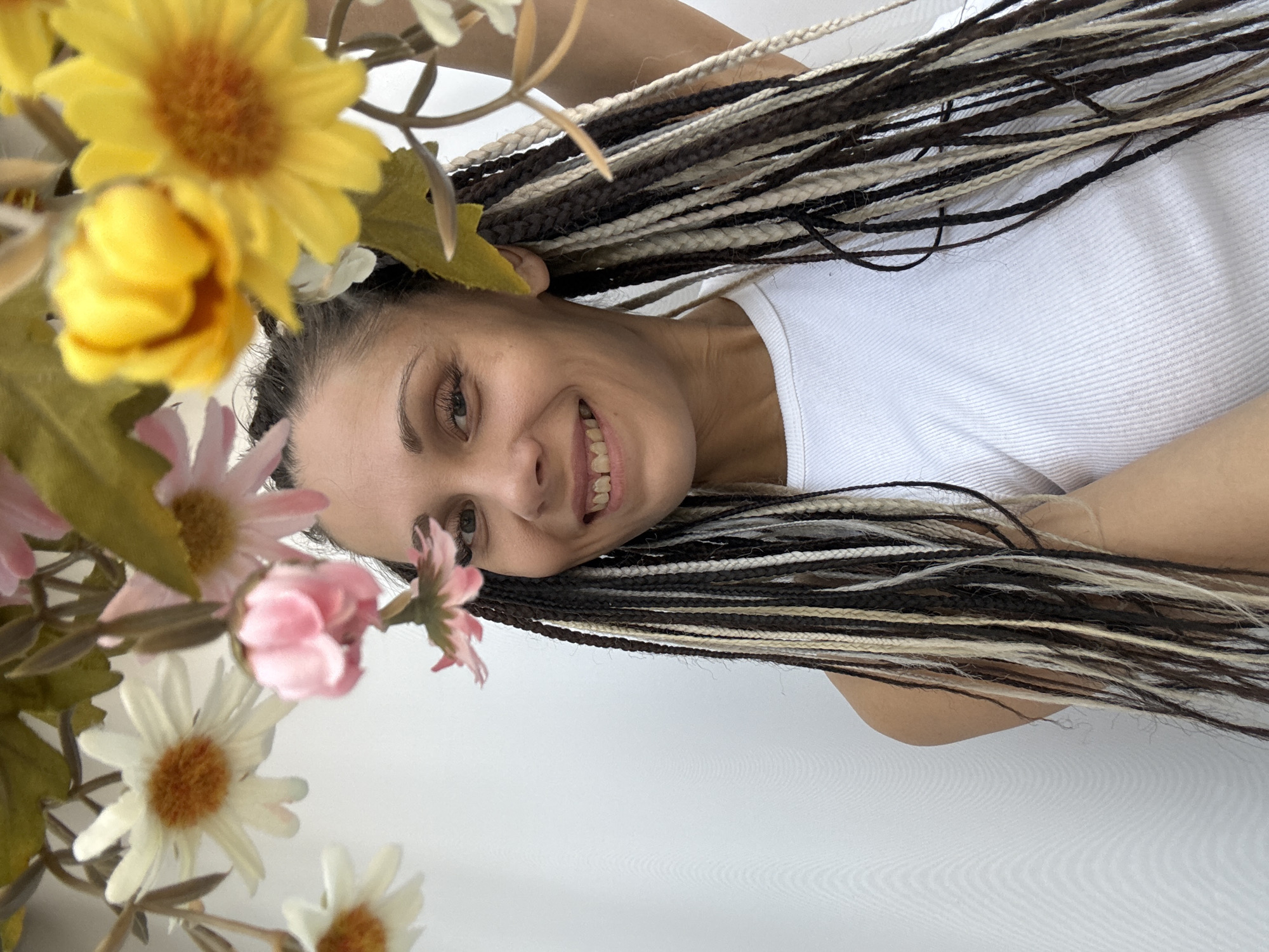 A woman with braided black and blonde hair is smiling while wearing a white tank top. She is surrounded by artificial yellow and pink flowers. The location appears to be indoors with a plain white wall. The city and precise location are unknown.