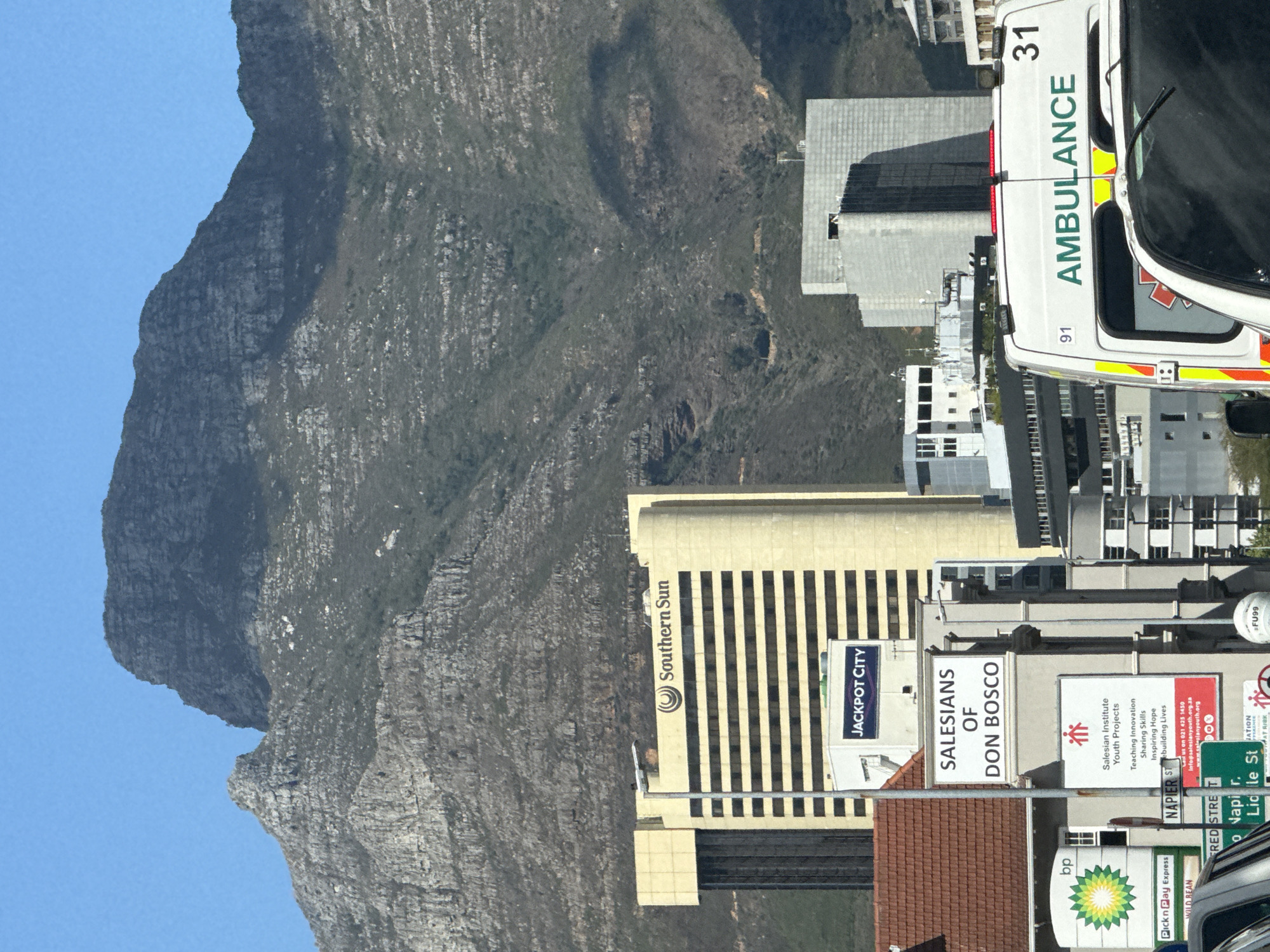 This image captures a vibrant urban scene in Cape Town, South Africa, set against the dramatic backdrop of a large, rocky mountain, strongly resembling Table Mountain, under a clear blue sky.
In the mid-ground, several multi-story buildings stand out. A prominent yellow building with vertical dark stripes displays the "Southern Sun" logo and a "JACKPOT CITY" sign. Adjacent to it, another building features signage for "SALESIANS OF DON BOSCO."
On the street level in the foreground, an ambulance, primarily white with green and yellow reflective markings, is partially visible. The word "AMBULANCE" is clearly written in green capital letters on its side, accompanied by the numbers "31" and "91."
Below the Salesians building, a large sign details the mission of the "Salesian Institute Youth Projects," listing "Teaching Innovation," "Sharing Skills," "Inspiring Hope," and "building Lives." Contact information, including "021 425 1450" and "info@salesianyouth.org.za," is also visible. Street signs indicating "NAPIER ST" and "FRED STRE T" are present. Further commercial establishments are suggested by a "bp" logo, a "Pick n Pay Express" store, and "WILD BEAN" signage.
The time of day is bright daylight, indicated by the clear blue sky and strong illumination. The overall scene is static, capturing a moment in a bustling city environment.