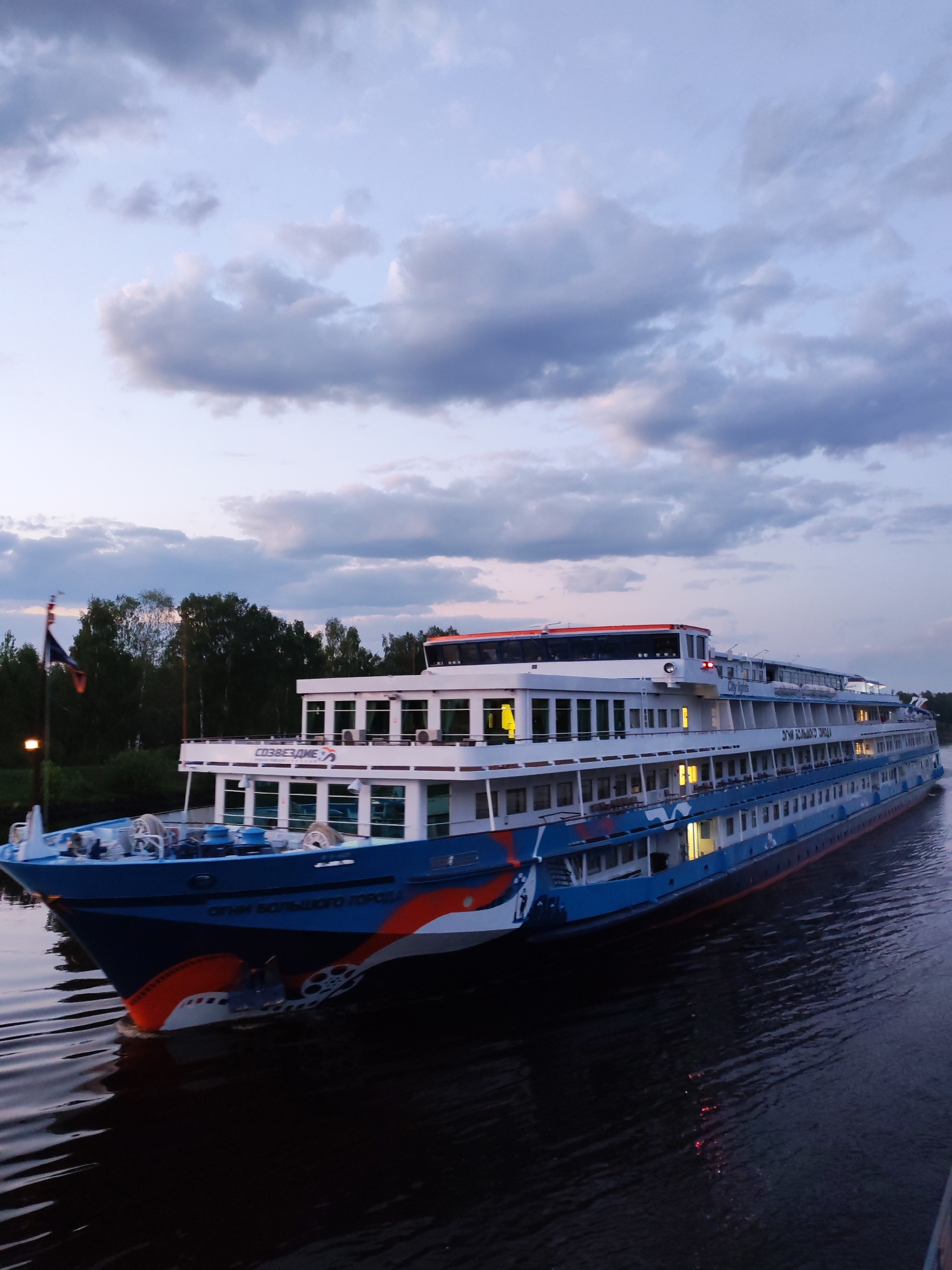 A long passenger ship, "Ogni Bolshogo Goroda," is traveling on a body of water, likely the Moscow Canal near the city of Zaprudnya, Russia, based on the ship's route. The ship is blue and white with some red and orange accents on the bow. The sky is cloudy and appears to be dusk or dawn. The shore is wooded with deciduous trees. A flagpole with a flag on it is on the left.