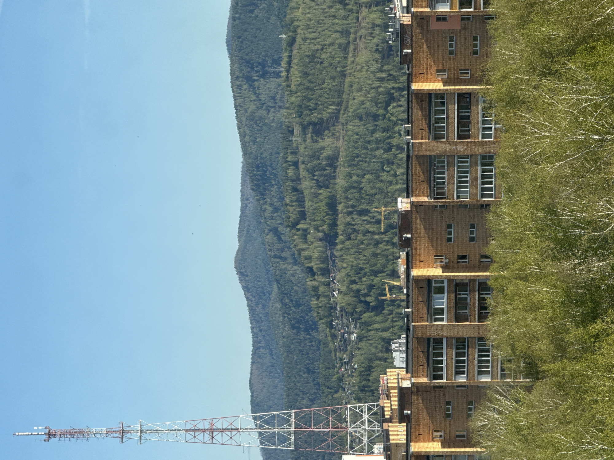 The image shows a tall, brick residential building in Krasnoyarsk, Russia. The building has multiple floors with rows of windows. Dense green trees are adjacent to the building. In the background, there is a mountain range covered in forest, and the bright blue sky completes the landscape. A lattice telecommunications tower stands in the foreground, extending towards the sky. A construction crane is also visible next to the building.