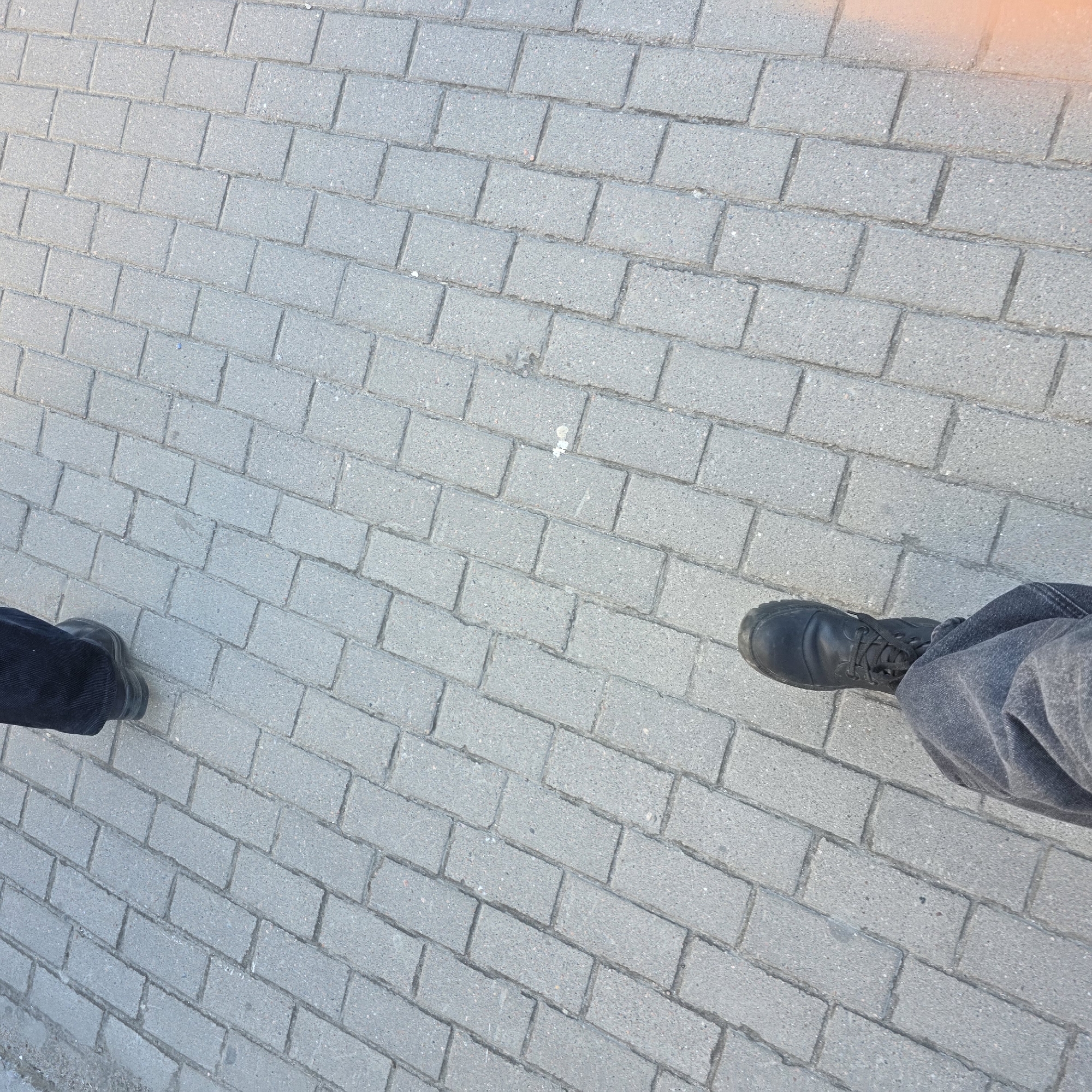 The image shows a paved area, possibly a sidewalk or pedestrian zone, constructed from grey rectangular paving stones arranged in a running bond pattern. Two feet, presumably belonging to the photographer, are visible. The foot on the left is wearing a dark, possibly black, shoe or boot, while the foot on the right is wearing a black boot and dark denim jeans. The image is angled, giving a perspective view of the ground. The exact location cannot be determined from the image alone.