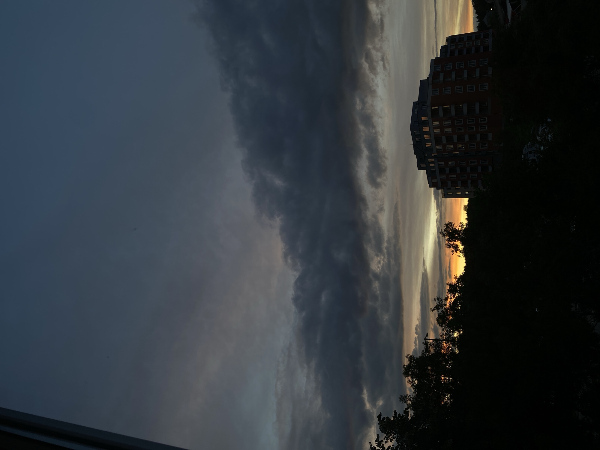 This is a vertical, medium-shot photograph of a city skyline at dusk. The sky dominates the upper two-thirds of the image, filled with layers of dark, heavy clouds that are tinged with the warm hues of sunset in the distance. A large, multi-story apartment building with a reddish-orange facade and numerous windows, some illuminated, is silhouetted against the sky and trees in the middle ground. Dark foliage, likely trees, fills the lower third of the image, obscuring the ground level and any details below the building. The overall mood is somber and dramatic, with the fading light creating a sense of deep shadow and muted colors.