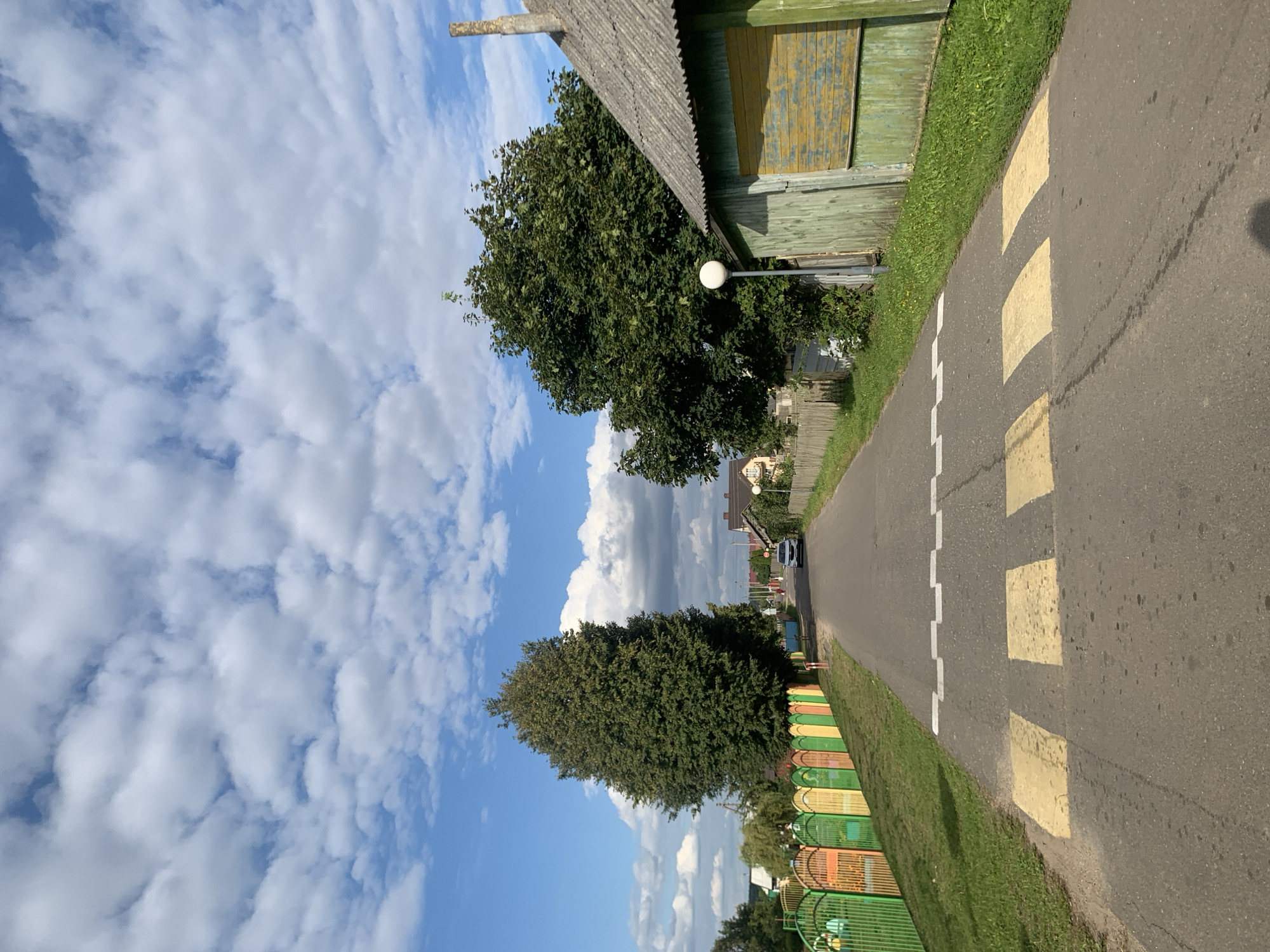 In Kapyĺ, Belarus, a paved road stretches through a residential area under a bright blue sky dotted with fluffy white clouds. The road features a pedestrian crossing with yellow stripes and a speed bump marked with white zigzag lines. On the left side of the road, a vibrant fence with multicolored panels in green, yellow, orange, and red borders a grassy area. Tall, leafy trees stand behind the fence. Further down the road, a white car is visible. On the right side of the road, a rustic wooden building with a weathered green facade and peeling yellow shutters is situated next to a tall, dark green tree. A street lamp stands in front of the building. Wooden fences line the properties along the right side of the road. Houses with pitched roofs are visible in the background.