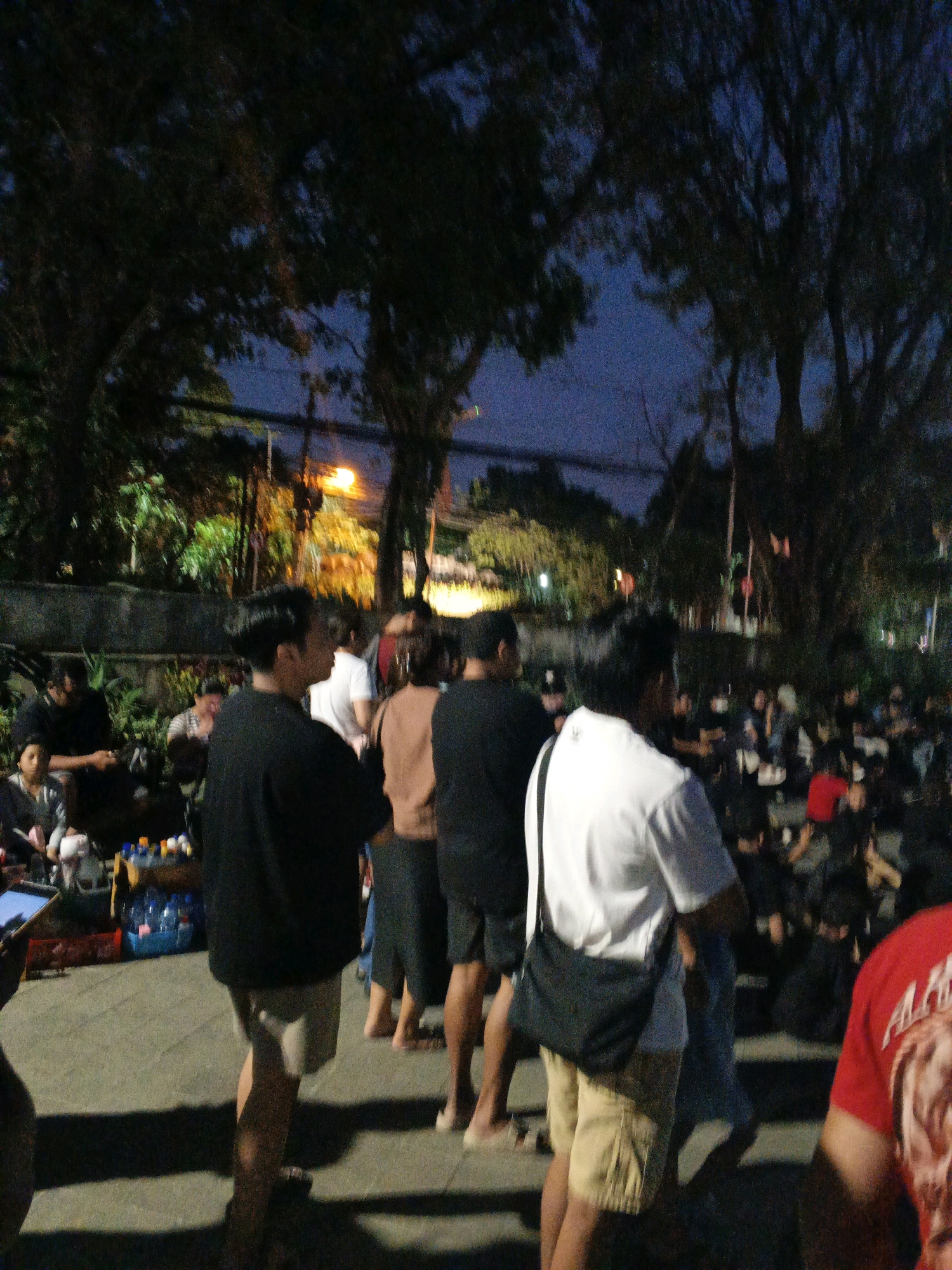 The image depicts an outdoor gathering at night, likely in Denpasar, Indonesia, with a group of people standing and sitting. The environment features trees and artificial lighting, suggesting a park or public space. Several individuals are visible, with a focus on a man in the foreground wearing a white t-shirt and a black bag slung across his shoulder. He is facing away from the camera. Other people are seen in various poses, some seated on the ground, others standing in a line. Objects like bottles of water and a red crate are present, indicating a casual, possibly informal event. The overall atmosphere is dimly lit, with the dark sky and tree foliage creating a subdued ambiance.