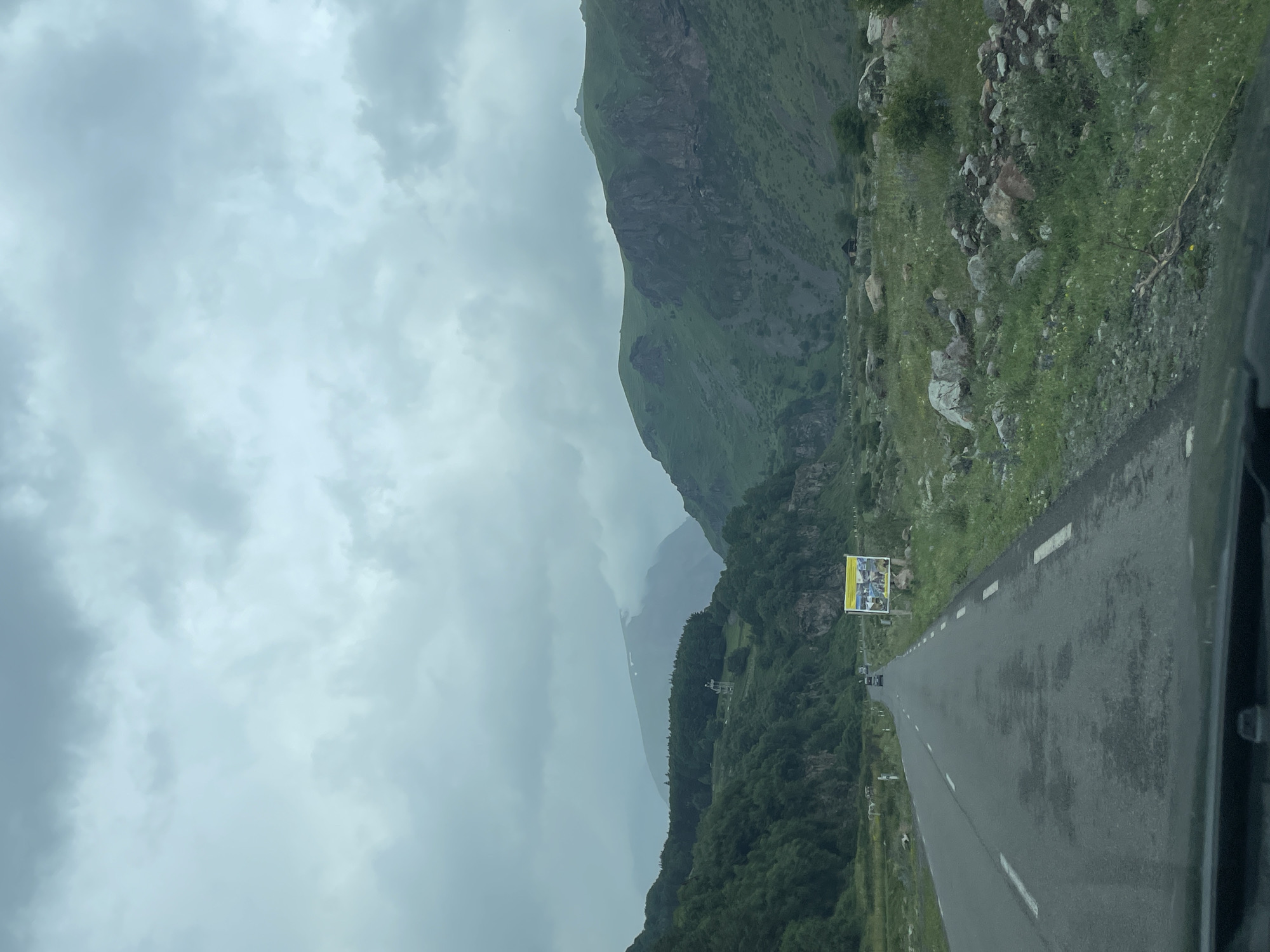 An overcast, cloudy day is captured in this outdoor, eye-level, full-shot photograph, looking down a winding asphalt road through a mountain valley. The road is lined with white dashed lines on the right and a solid white line on the left, and appears to be in fair condition with some darker patches of wear. A few vehicles are visible on the road in the distance, heading away from the viewer. To the right of the road, there is a large billboard with yellow and white signage. The landscape on either side of the road is rugged and mountainous, with steep, green, rocky slopes rising sharply. Dense green trees cover the lower portions of the mountains. The valley floor is covered with rocks and sparse green vegetation. The sky is filled with heavy, gray clouds, suggesting impending rain or a generally damp day. In the background, a prominent mountain peak is visible, partially obscured by clouds. The scene appears to be in Gergeti, Georgia, known for its dramatic mountain landscapes.