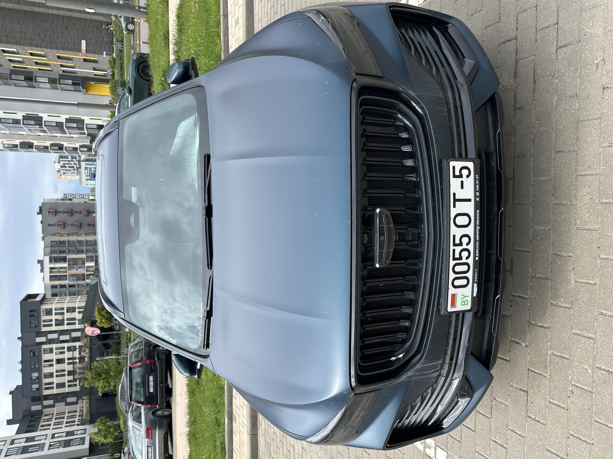The image shows the front of a blue Geely car parked on a paved area, likely in Бараўлянскі сельскі Савет, Belarus, as indicated by the Belarusian license plate. The license plate number is BY 0055 OT-5. The grill of the car is prominently visible with the Geely logo. In the background, there are several residential buildings and some greenery, indicating an urban or suburban setting. Several other parked vehicles are visible in the background as well. The sky appears overcast.