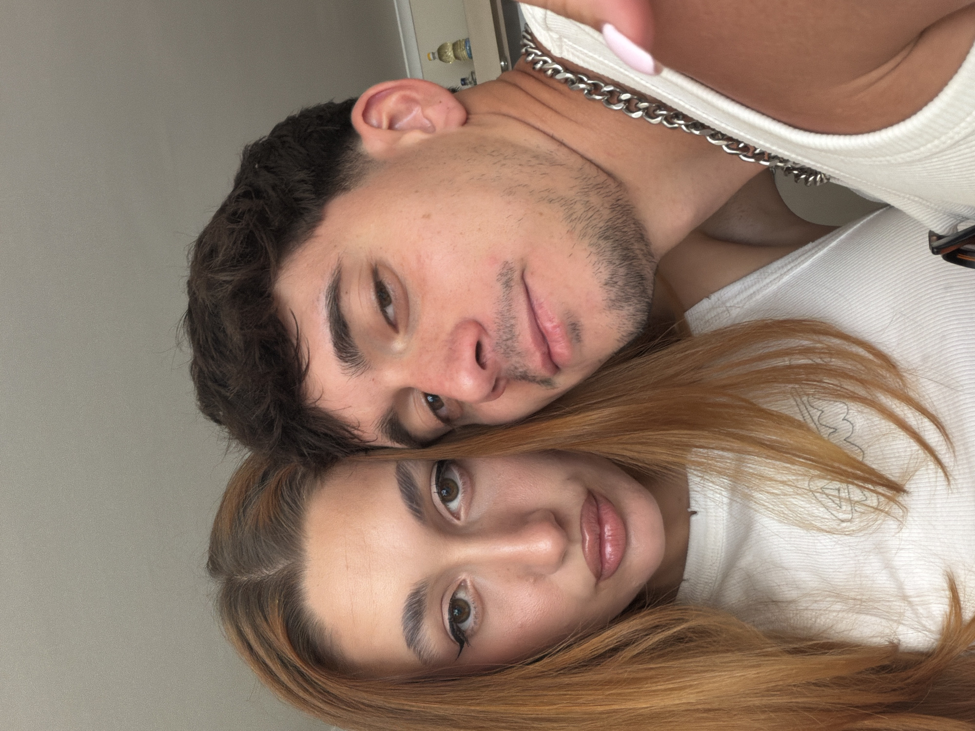 This image, reportedly taken in the city of Sofia, Bulgaria, captures a close-up selfie of two individuals, a female and a male, posing indoors.
The female occupies the left and central portion of the frame, directly facing the camera with a composed, slightly pouting expression. She has long, straight, reddish-brown hair parted down the middle, with visibly darker roots. Her eyes are brown, accentuated with dark cat-eye eyeliner, and her eyebrows are thick and well-defined. Her lips are full and appear glossy with a pink tint. She wears a white ribbed tank top, featuring a subtle, light-colored abstract graphic (resembling "WM" or a wave) on the chest.
To her right, the male subject leans into the frame, his face partially obscuring the right side of her head. He has short, dark brown, wavy hair and a faint stubble beard on his jawline and mustache area. His brown eyes are directed slightly off-camera to his left. He also wears a white ribbed tank top and a thick silver chain necklace. A finger with light pink nail polish is visible in the lower right corner of the frame, adjacent to the male's shoulder.
The background is a plain, light grey wall or ceiling. In the far upper right, a blurry shelf is visible, holding two small bottles, one yellow and one clear, suggesting a domestic interior.