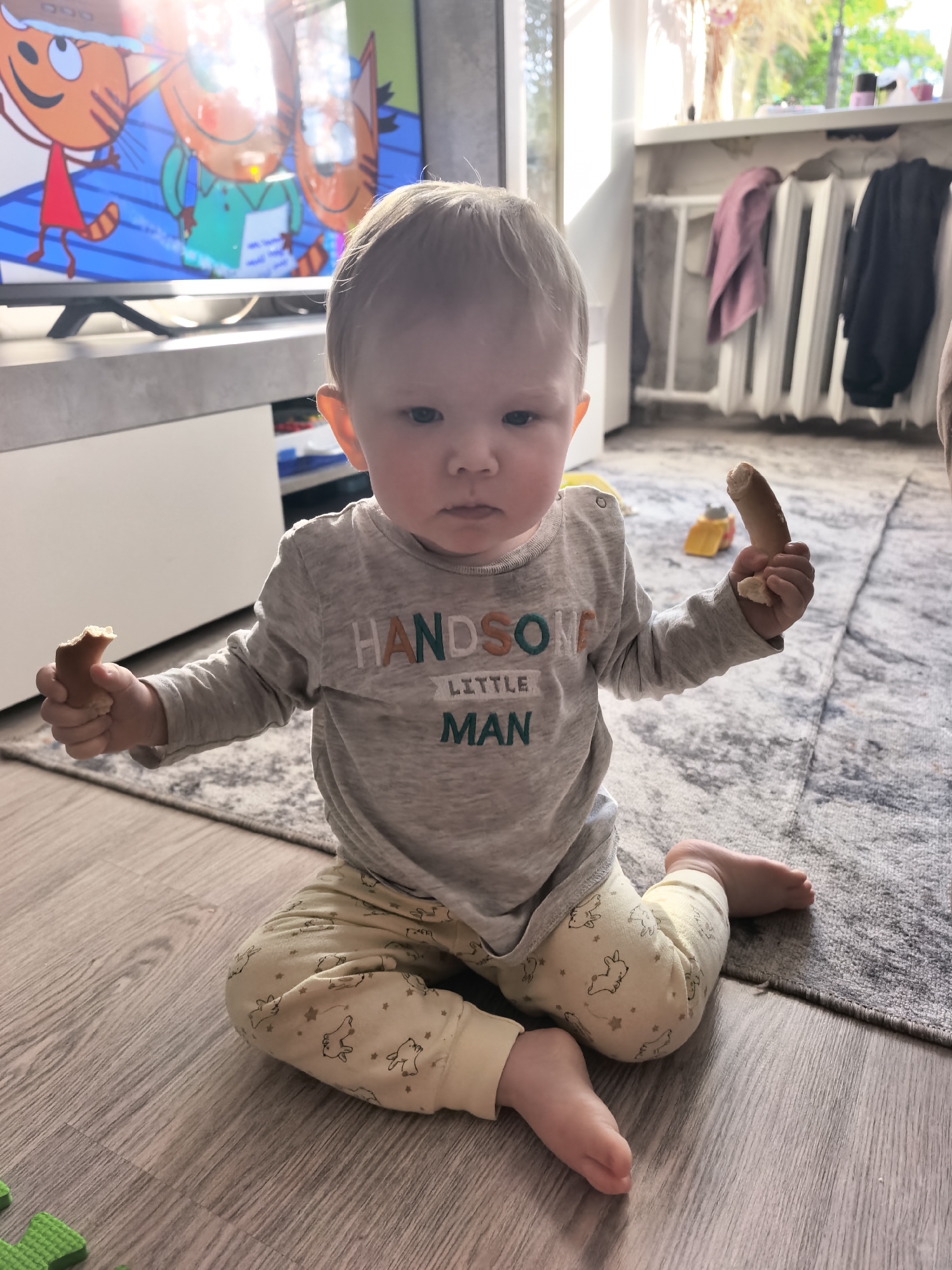 An overhead, eye-level, full-frame indoor photograph taken in Sterlitamak, Russia, depicts a fair-skinned toddler, approximately 12-18 months old, with light hair and blue eyes, sitting cross-legged on a light grey wooden floor. The child wears a long-sleeved grey shirt with "HANDSOME LITTLE MAN" printed on the chest in orange, green, and white letters, and light yellow pants adorned with a pattern of bunnies and stars. The child holds a piece of a light-brown, elongated food item (possibly a breadstick or bagel) in each hand, looking directly at the viewer with a neutral expression.
To the left of the child, a green foam puzzle mat piece is visible. Behind the child, on a grey patterned area rug, a small yellow toy vehicle (resembling a dump truck) is present. In the background, to the left, a flat-screen television displays an animated program featuring three anthropomorphic orange cats, with bright sunlight reflecting off the screen from a window. To the right of the child, a bright window reveals green outdoor foliage and sunlight, below which a white multi-section radiator is visible with a purple cloth draped over it. Adjacent to the radiator, dark garments are hanging. The room is brightly lit by natural sunlight.
