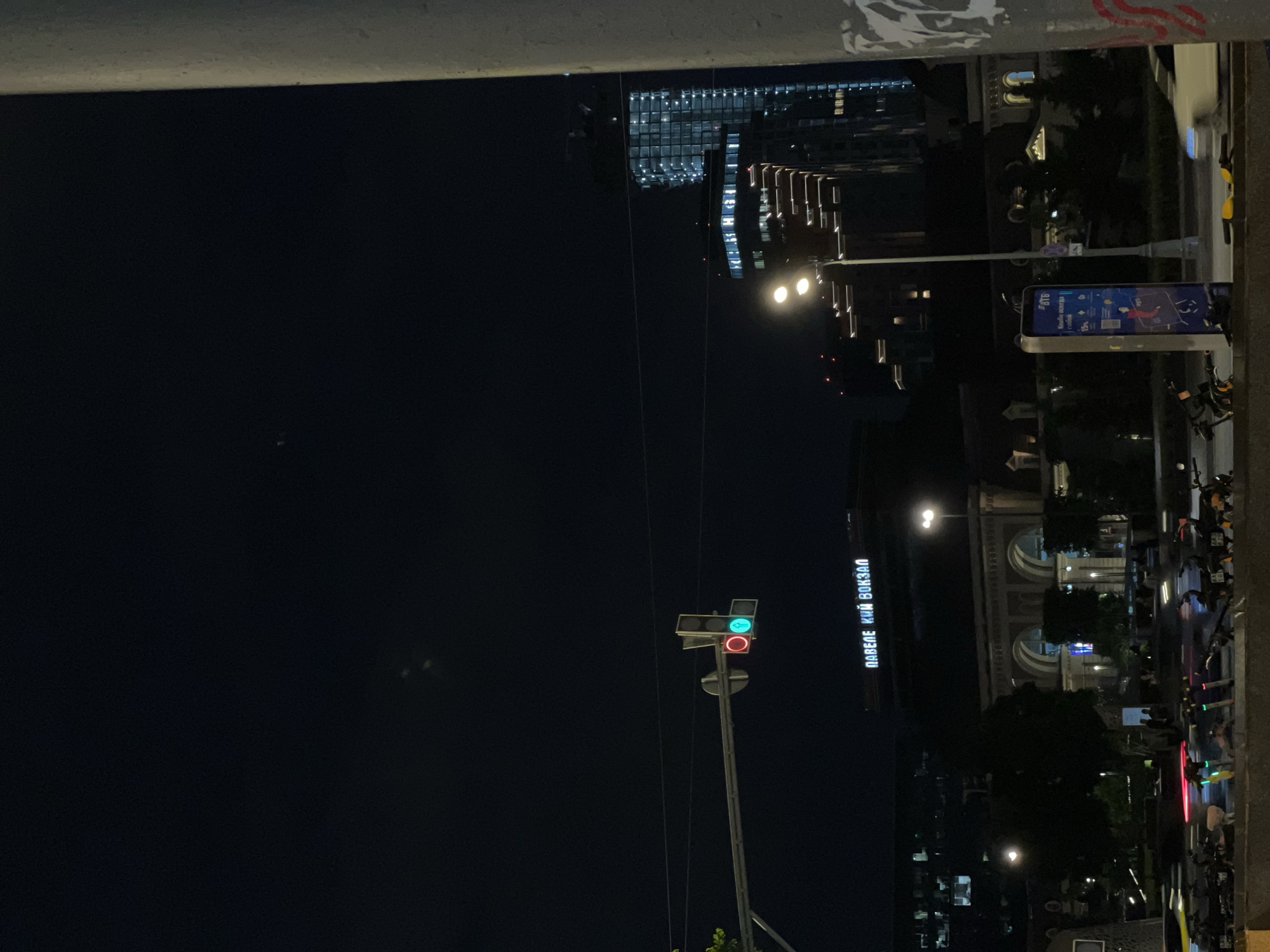 The image shows a night scene in an urban area, possibly near a train station. There is a traffic light with a green light illuminated on top of a red light, and a sign that reads "ПАВЕЛЕ ЦКИИ ВОКЗАЛ", indicating the location is likely near the Paveletsky railway station. A modern, high-rise building with lighted windows is in the background. There are streetlights and a digital advertising panel along the roadside. Several bicycles and scooters are parked near the advertising panel. The environment appears to be a busy city street at night.