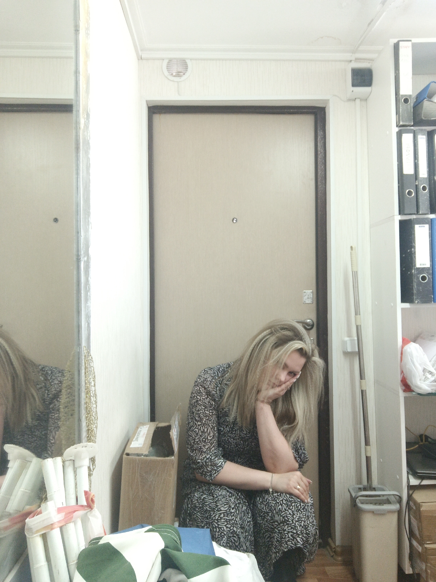 A woman with long, light-blonde hair is seated on the floor in what appears to be an interior entryway or hallway in Noviy, Russia. She wears a long-sleeved dress with a black and white abstract pattern. Her posture suggests weariness or contemplation; her right elbow rests on her knee, supporting her head with her hand, while her left hand rests on her left knee. She has a ring on her right hand and a bracelet on her left wrist, and her expression seems somber or tired as she looks towards the viewer.
She is leaning against a light-colored metal door with a dark brown frame, which features a peephole and a lock mechanism. To her left, a tall mirror reflects her upper body, hair, and dress, as well as some bundled white plastic rods tied with a red and white ribbon and a green and white checkered fabric on the floor. To her right, a white shelving unit is filled with several binders, predominantly black, with some visible text, including "STAFF" on one. A blue binder is also present.
Above the door, a circular ventilation grille is visible, and to the right, a small white box, possibly an alarm system or sensor, is mounted on the wall. A metallic mop leans against the wall next to the shelving unit, with a beige mop bucket below it. A partially open cardboard box is situated behind the woman, and another cardboard box is at the bottom right. The floor is made of light-colored wood. The overall setting appears to be a functional, utilitarian space, possibly an apartment or office corridor. There are no direct cues for the time of day or weather, as it's an indoor scene.