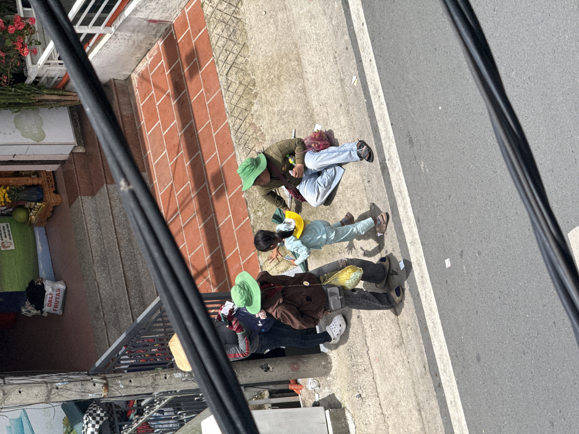 In Da Lat, Vietnam, two individuals and a child are present on a sidewalk next to a street. The sidewalk is made of concrete, with a white stripe marking the edge of the road. To the left of the sidewalk is a building with a concrete pillar and a metal railing. The building has stairs leading up to it, and the ground level appears to be a shop. The people are dressed in casual clothing. One person, wearing a brown jacket and a green hat, is seated on the sidewalk. Another person, wearing a gray jacket and a green hat, is standing and holding a yellow bag. The child, wearing a light blue outfit and a yellow hat, is standing between them. The scene appears to be a street-level view, possibly from a higher vantage point, as evidenced by the black cables crossing the foreground. The environment suggests a typical urban setting in Da Lat, with a mix of commercial and residential elements.