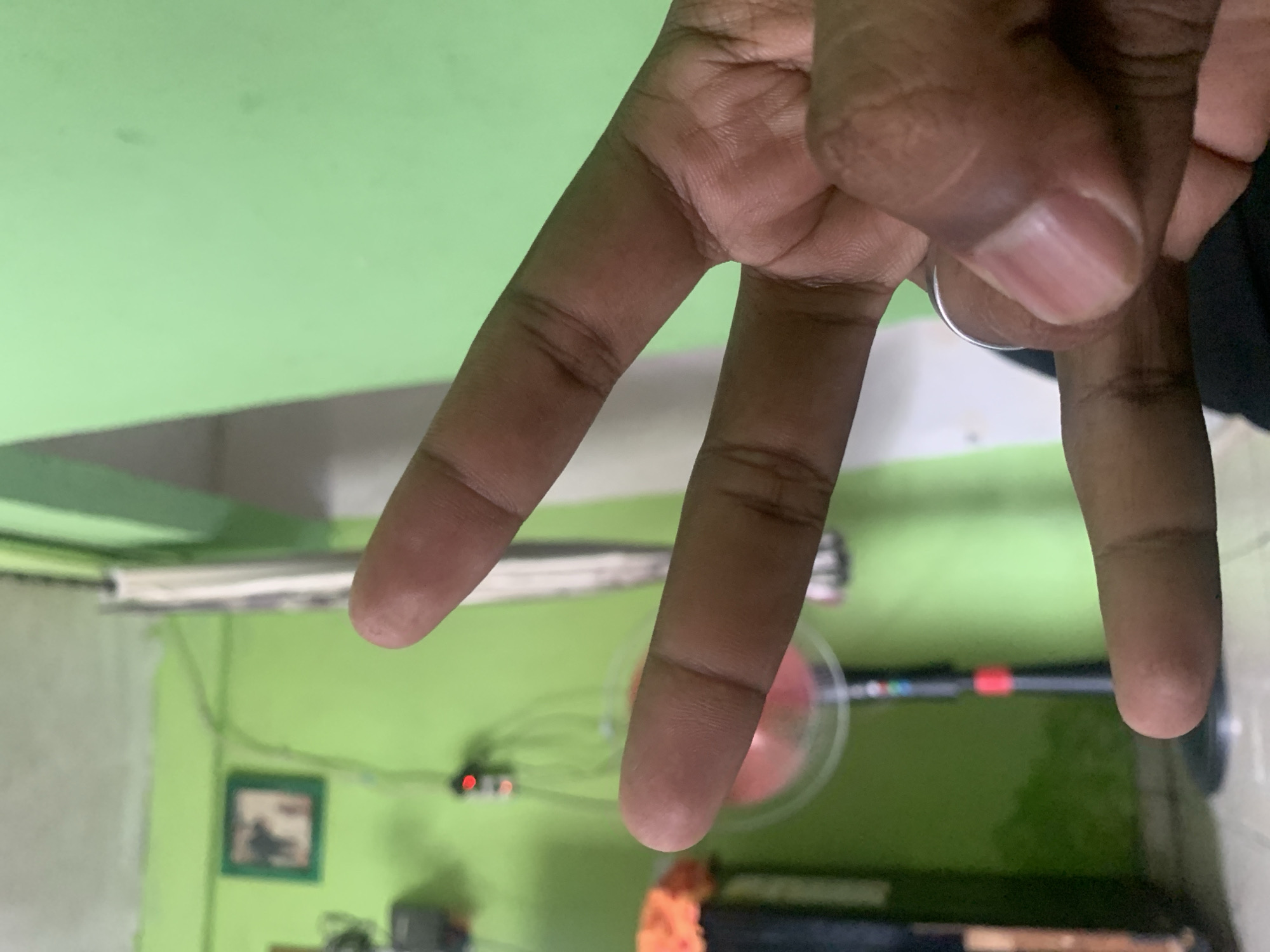 A close-up shot shows a person's hand holding up three fingers, forming the number three. The hand is in the foreground, with a green wall in the background. A blurry image of a fan can be seen in the background, and a framed picture is hanging on the wall. The scene is indoors, likely in a residential setting. The image does not provide any specific location within the city of Palembang, Indonesia.