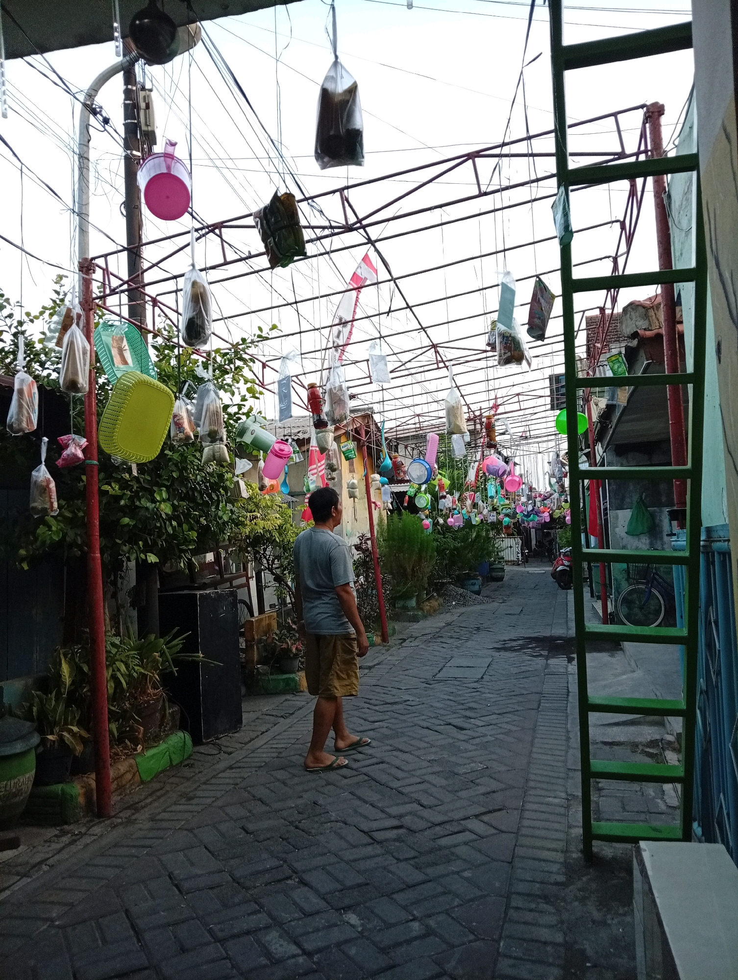 The image shows a narrow, paved alleyway in Surabaya, Indonesia, lined with buildings and greenery. A man in a gray t-shirt and khaki shorts stands in the middle of the alley, facing away from the camera. The alley is decorated with numerous items suspended from overhead wires and a metal frame, including plastic bags containing objects, colorful buckets, baskets, and other household items. The overall impression is of a community garden or a creative display of repurposed materials. The sky is overcast, suggesting it may be late afternoon or early morning.
