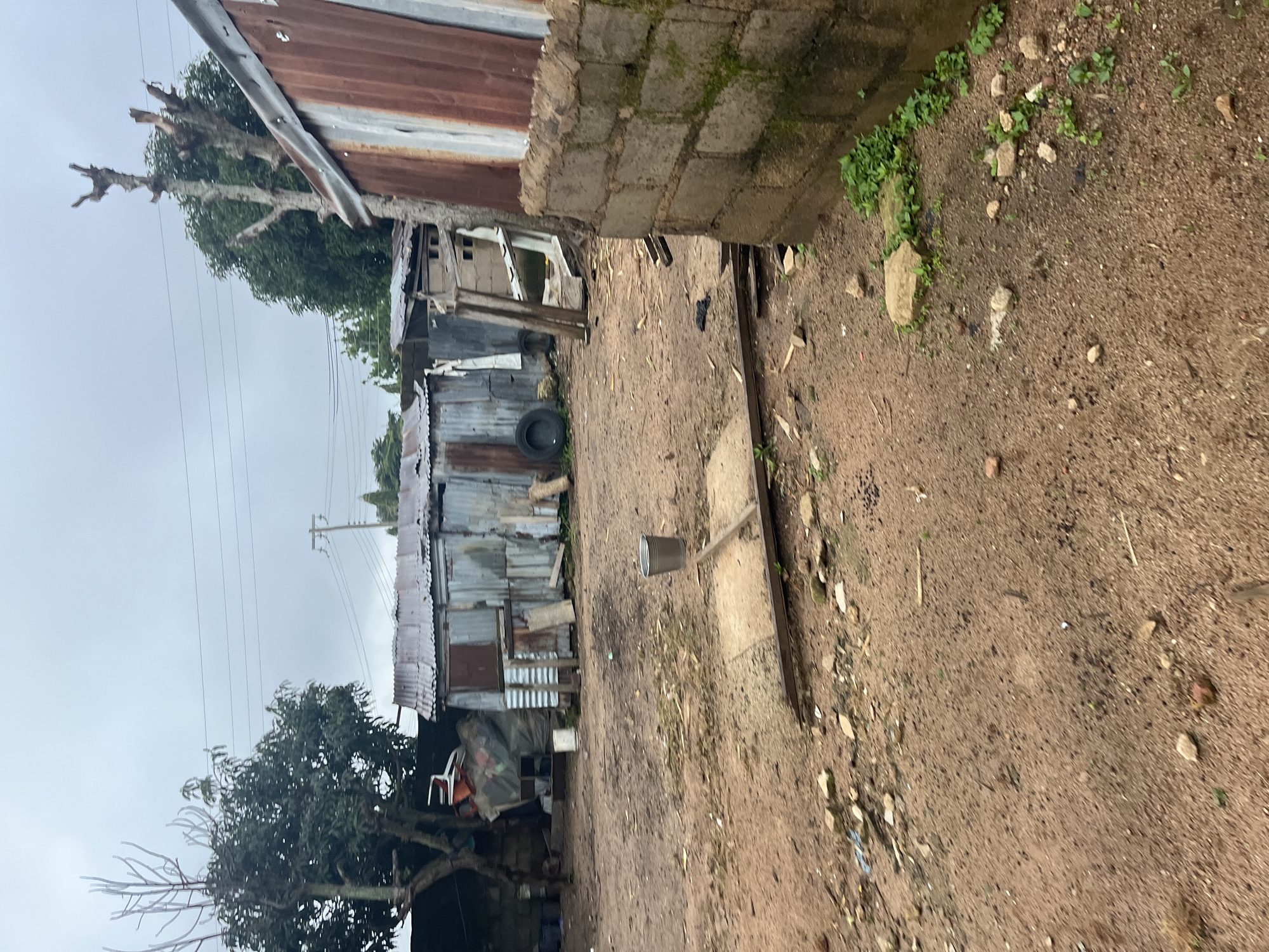 The image depicts an outdoor scene in a densely populated area, possibly a settlement within the city of Jos, Nigeria. The ground is a mix of dry, dusty earth and scattered debris, including rocks, twigs, and small pieces of plastic. Several makeshift structures, constructed primarily from corrugated metal sheeting, are visible. These structures appear weathered and in disrepair, with some showing signs of rust and damage.
In the foreground, a gray metal bucket sits on a concrete slab. To the right, a partially constructed wall made of concrete blocks is present, with moss or algae growing on its surface. In the background, vegetation is sparse, with a few trees, some of which are bare and appear to have been pruned. Overhead, power lines stretch across the overcast sky. The overall impression is one of a utilitarian and somewhat neglected environment.
