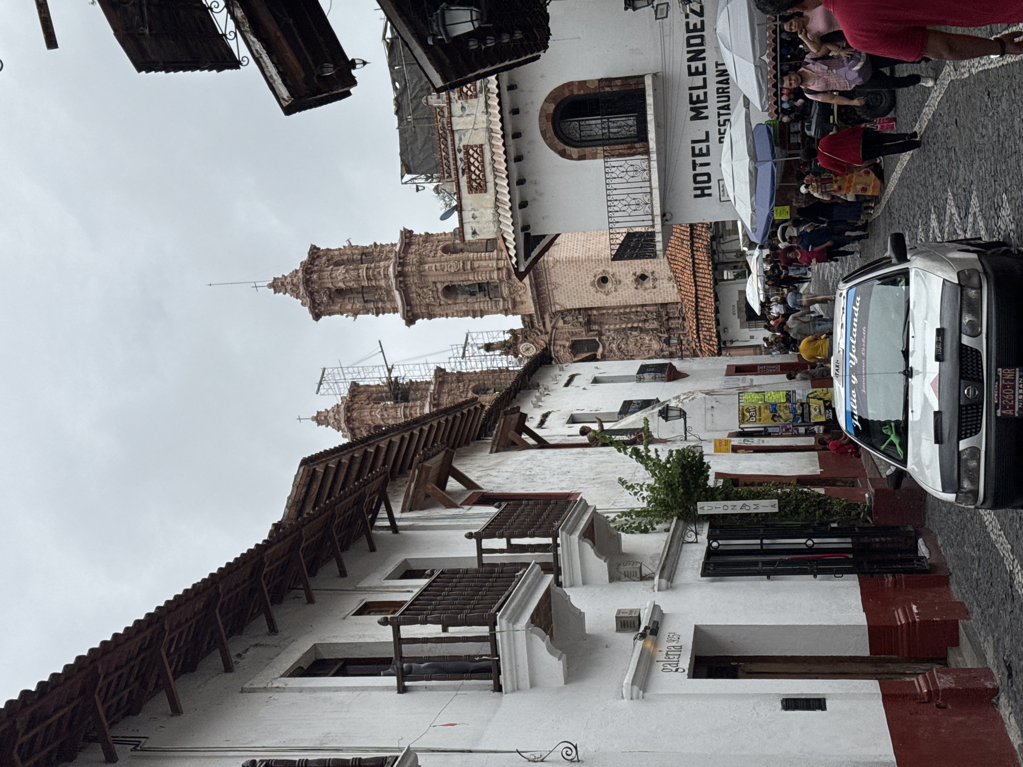 This detailed image captures a bustling cobblestone street scene in Taxco, Mexico, characterized by its traditional architecture under an overcast sky.
In the background, two prominent reddish-brown baroque church towers, likely belonging to the Santa Prisca de Taxco church, rise above the surrounding buildings; the left tower features visible scaffolding. The street, paved with dark cobblestones, curves gently to the right as it recedes into the distance.
On the left side of the street, a line of multi-story buildings with white stucco walls and dark red bases features dark brown wooden balconies and window frames. The roofs are composed of red tiles. A building in the foreground on the left has a recessed entrance marked "galeria 925" and a dark metal gate adjacent to it. Further along, a small tree grows against a building with a sign that reads "AUTONOMÍA." One balcony on the left features a dark, human-like figure, possibly a mannequin or sculpture.
On the right side of the street, similar white buildings with red tile roofs are present. A large sign for "HOTEL MELENDEZ RESTAURANT" is prominently displayed on a building further down the street. Below this sign, several white and blue market umbrellas are set up, sheltering pedestrians and possibly vendors. A person is visible leaning over a balcony on a building on the right, midway down the street.
Numerous pedestrians populate the street, walking in various directions, primarily concentrated on the right side near the market stalls. Their attire is casual, including individuals in yellow and red shirts.
A white Nissan Tsuru sedan, identifiable as a taxi by a roof banner that reads "Julio y Yolanda," is positioned prominently in the foreground, facing the viewer. A small green frog toy (Kermit) rests on its dashboard. The license plate "A-260-FNR" is visible. Partial view of a four-wheeled vehicle, possibly an ATV, is seen behind the market umbrellas on the right.