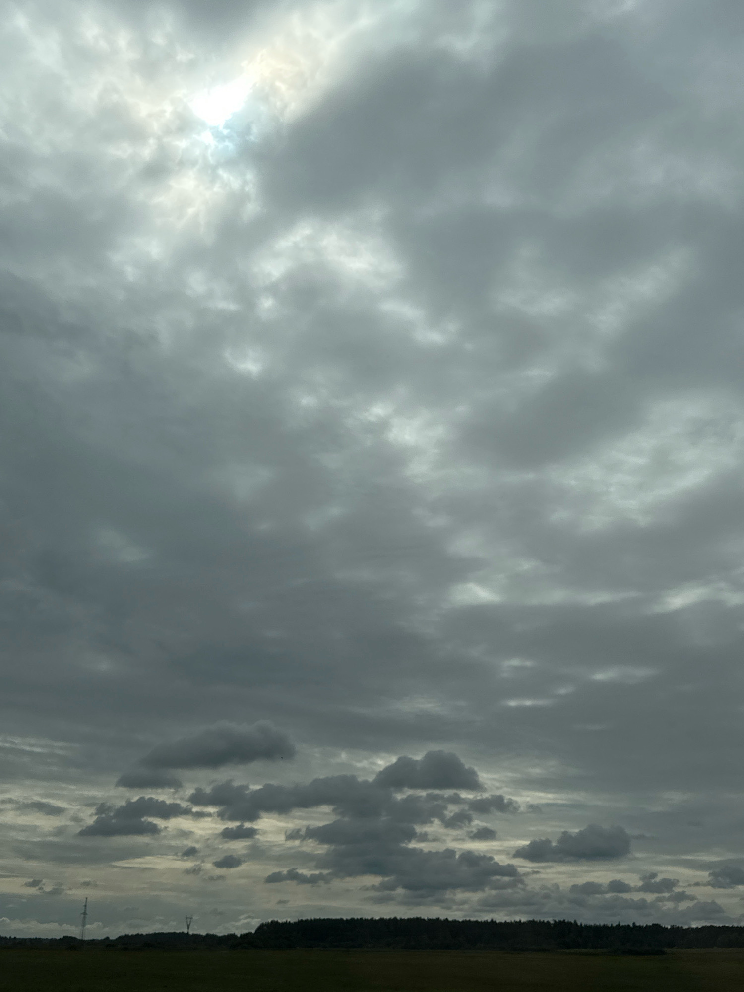 The image depicts a vast, overcast sky with scattered patches of sunlight breaking through. The clouds are a mixture of grey and white, with varying textures and densities. Below the sky, a dark, flat landscape stretches across the horizon, characterized by a line of trees and a field. Two utility poles are visible in the lower left and center of the frame, suggesting an open, possibly rural area. The lighting suggests either early morning or late afternoon, or a generally cloudy day. The scene is devoid of human presence or any overt activity, presenting a tranquil, natural environment. The location is specified as being within the village of Garbatsevichsky Selsoviet, Belarus.