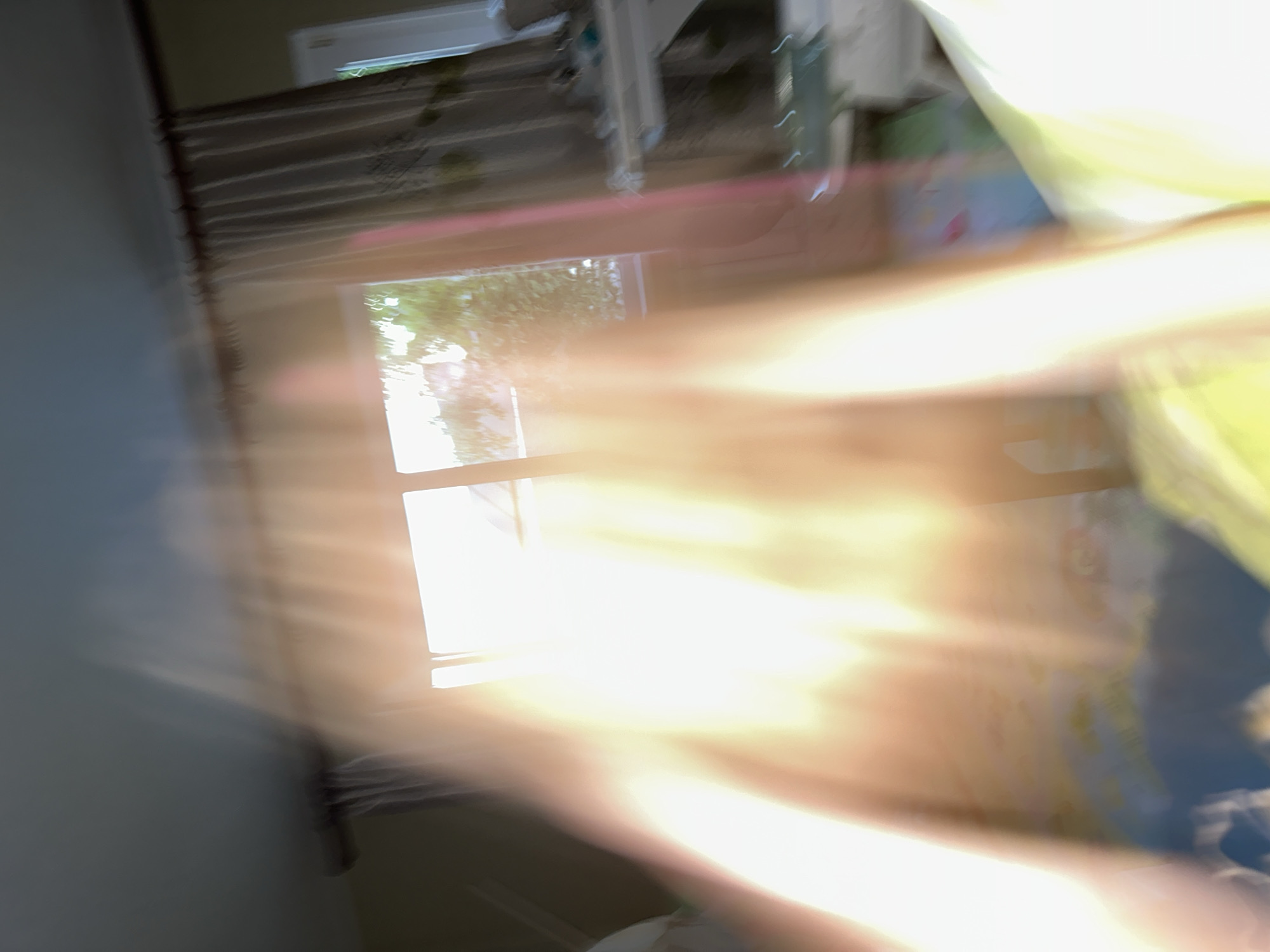 The image depicts a blurred interior scene, likely a bedroom, with a window that allows natural light to enter. The light creates a bright, washed-out effect, obscuring many details. A curtain is visible on the right side of the window, and what appears to be a dresser or shelf unit is partially visible. The foreground is dominated by a bright, glowing light source, possibly a flash or direct sunlight, which further blurs the image. The overall impression is one of motion blur and overexposure, making it impossible to identify specific objects or people with certainty or to determine a location within Nizhny Novgorod, Russia.