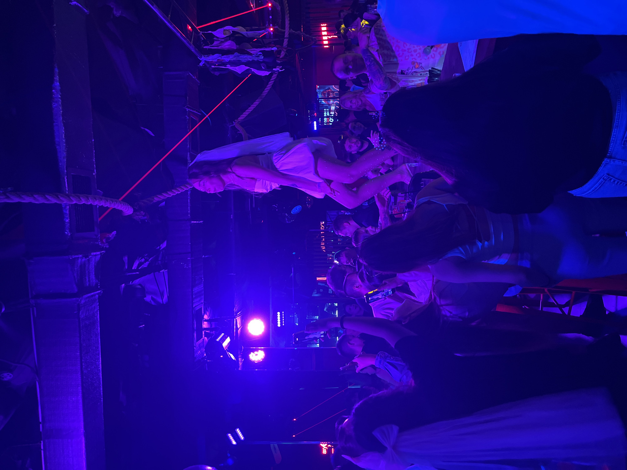 The image depicts an indoor scene at a nightclub or bar in Naberezhnye Chelny, Russia, bathed in vibrant purple and blue lighting. In the center, a young woman wearing a short white dress and a veil is standing on a bar top, seemingly performing or posing. She is holding onto a thick rope hanging from the ceiling. Below her, a crowd of people are gathered, many of them holding up their phones to capture the moment. Some individuals are dancing or socializing, while others appear to be watching the performance. The environment is dimly lit, with neon lights and stage lighting contributing to the energetic atmosphere. In the background, there are various bar elements, including shelves with bottles and a screen displaying text. The overall impression is one of a lively celebration or event, likely a bachelorette party given the bride-like attire of the performer.