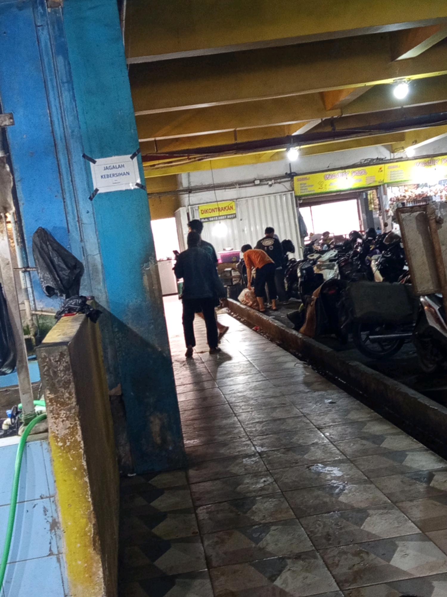 The image depicts an indoor market scene, likely in Karawang, Indonesia, characterized by its tiled floor and overhead lighting. A prominent blue pillar on the left side of the image features a sign that reads "Jagalah Kebersihan," which translates to "Maintain Cleanliness." In the background, several individuals are visible, with some appearing to be interacting with parked motorcycles. The overall environment suggests a bustling commercial space, possibly a parking area within or adjacent to a market. The lighting is a mix of natural light from an opening and artificial light from ceiling fixtures, creating a somewhat dim and possibly wet or reflective surface on the floor.