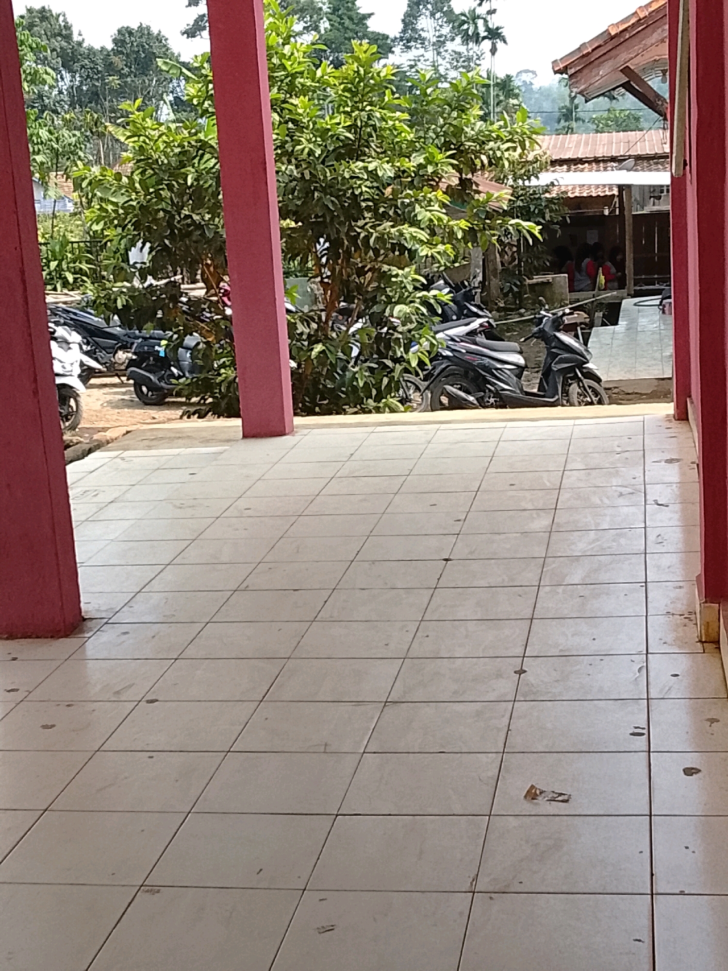The image depicts an outdoor scene in Tanjung Setia, Indonesia, likely in front of a building with a tiled walkway and red support pillars. Several motorcycles are parked on a dirt or gravel surface adjacent to the tiled area. In the background, there is lush green vegetation and trees, suggesting a tropical environment. Behind the trees, portions of buildings with tiled roofs are visible, and some people can be seen gathered under a covered area. The sky is overcast.