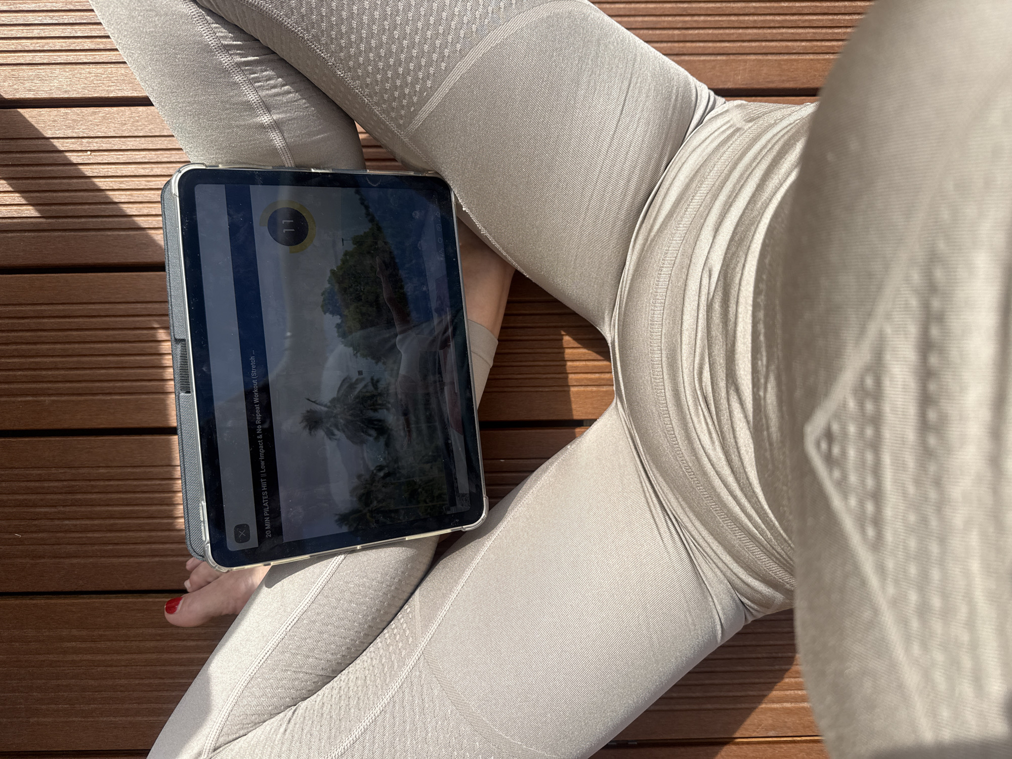 Here is a forensic description of the image:
The image shows a person sitting cross-legged on a wooden surface, likely a deck or patio. They are wearing grey leggings. An iPad rests on their legs, displaying a screen with a workout video titled "20 MIN PILATES HIIT || Low Impact & No Repeat Workout." A foot with red nail polish is visible. The scene appears to be outdoors, based on the lighting. Vila Nova de Gaia, Portugal is not discernible from the visual information provided.