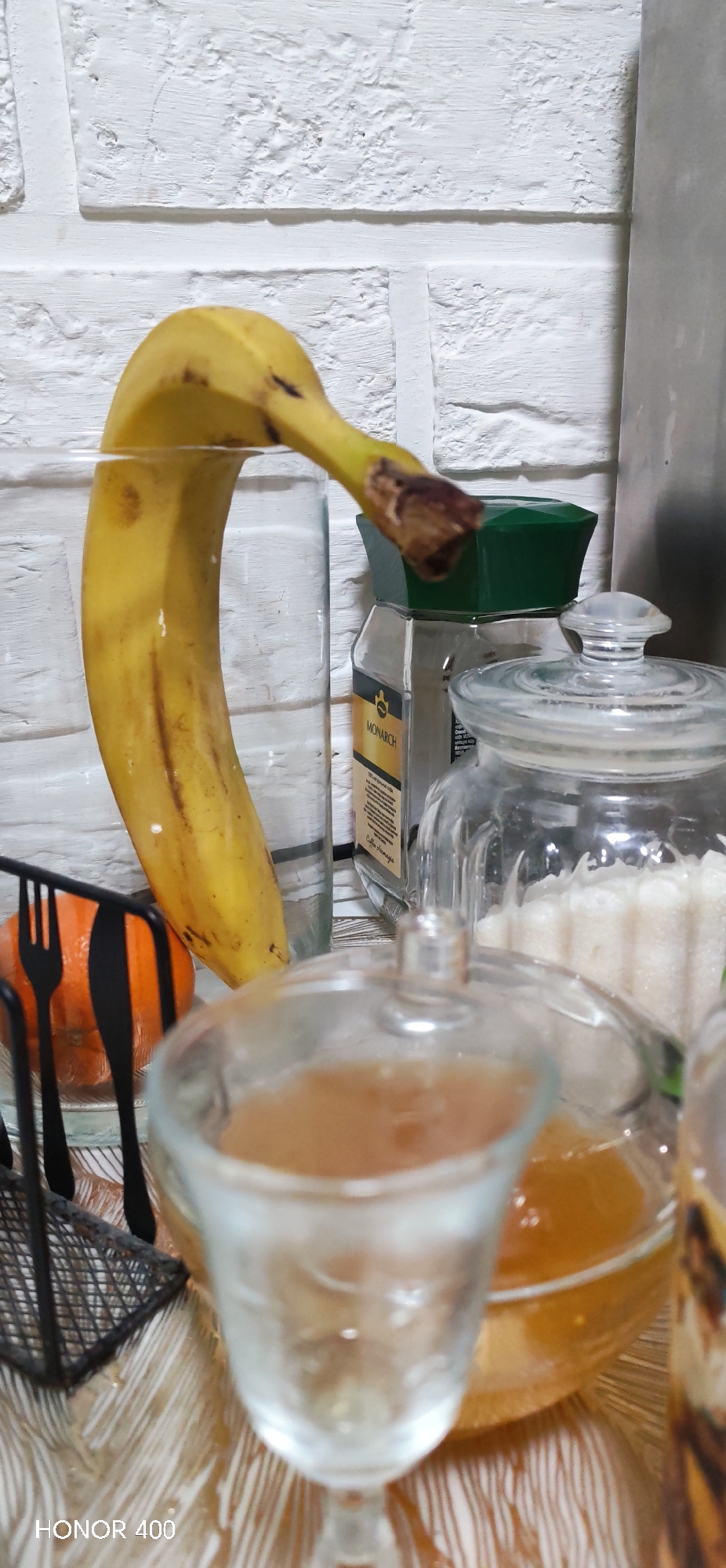 The image depicts a close-up, static indoor scene, likely a kitchen or dining area. In the foreground, a blurred clear glass stemmed goblet containing a light brown liquid is positioned slightly right of center. To its left, a black metal utensil holder, possibly a mesh design, contains dark-handled forks and knives. Partially obscured behind the utensil holder is an orange-colored spherical fruit. Dominating the central-left of the midground is a ripe yellow banana, curved and positioned vertically, with its lower section appearing to be within a large, clear, cylindrical glass container or vase, which is mostly transparent and indistinct. To the right of the banana, a clear glass jar with a dark green hexagonal lid is visible, bearing a label that reads "MONARCH" and "Coffee Amaga" in Cyrillic script. Further right, another clear glass jar with a clear glass domed lid contains a white, granular substance, consistent with sugar or salt. The background consists of a white wall featuring a textured, rectangular brick pattern. The objects rest on a surface patterned with alternating white and light brown or gold stripes. There are no people visible, and no active interactions are occurring. The specific location within Noviy, Russia, cannot be determined from the visual information presented.