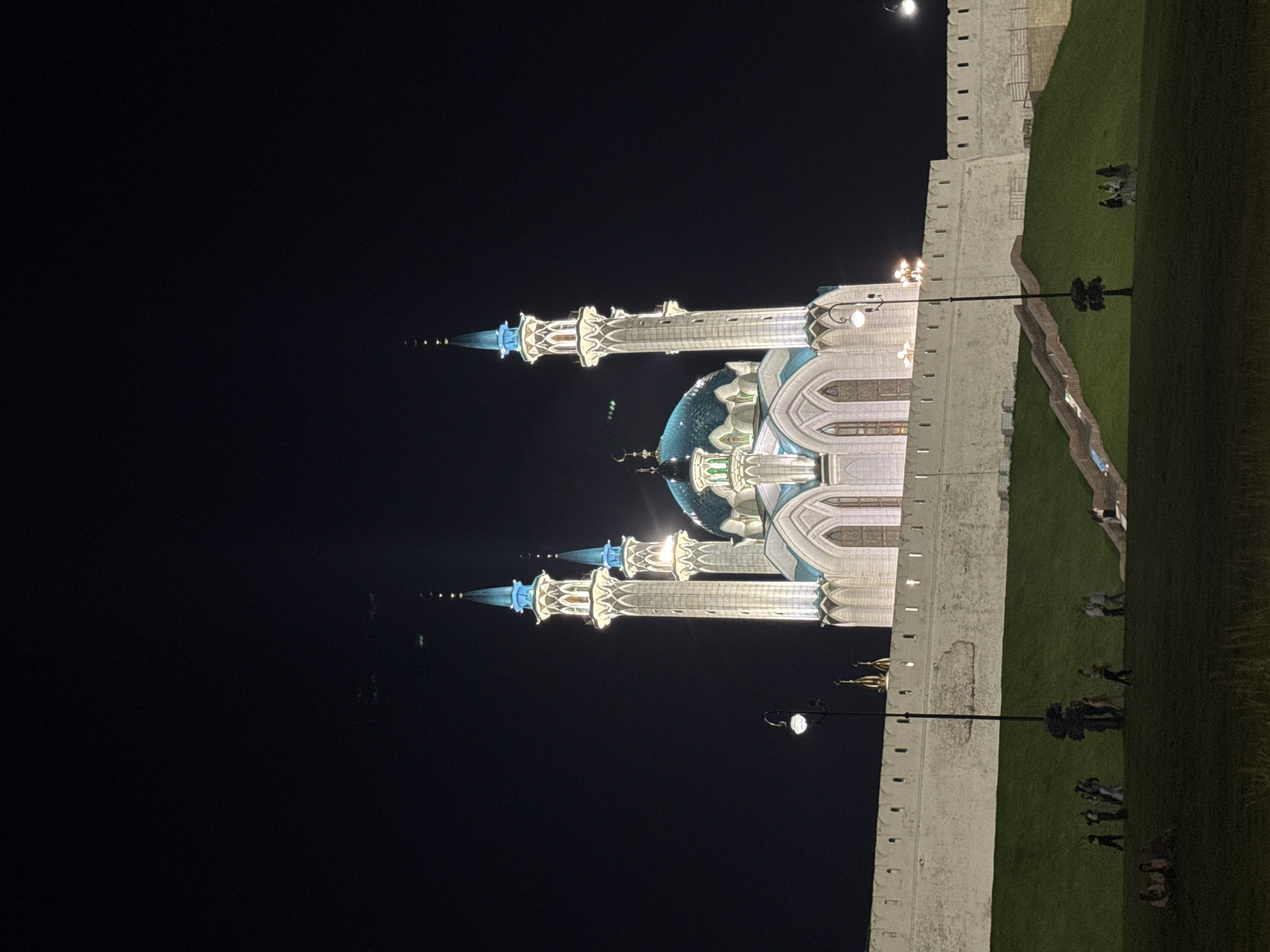 A night photograph captures the Kul Sharif Mosque in Kazan, Russia, illuminated against a dark sky. The mosque features white walls and several tall minarets topped with blue spires, each adorned with a crescent moon. A large turquoise dome is visible in the center of the structure. In the foreground, a grassy hill slopes upwards towards a white stone wall that surrounds the mosque complex. Several streetlights are positioned along the wall. A group of people are visible on the grassy area in front of the wall, some standing and some sitting. A set of stairs leads up the grassy slope towards the wall. The overall scene is dimly lit by the mosque's illuminations and the streetlights, creating a dramatic nighttime view.