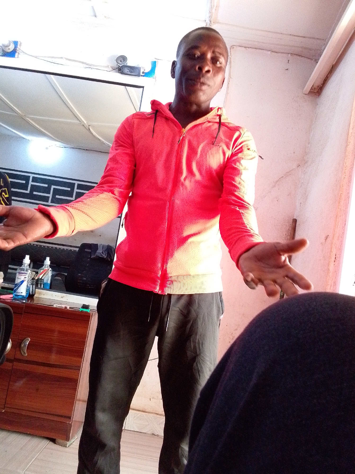 A dark-skinned man stands centrally, facing the viewer with an open, questioning or gesturing posture. He is wearing a bright, reddish-orange hooded zipper jacket and black athletic pants. His hands are open, palms slightly upward, extended out from his sides. His expression is neutral, with his mouth slightly ajar.
The setting appears to be an indoor space, possibly a casual business or personal area. To the left of the man, a dark brown wooden cabinet with two silver handles is visible. On its surface, several bottles are arranged, including two clear spray bottles, one containing a blue liquid. Text on one bottle is partially visible, reading "BARBER" and "AFRO". Another bottle seems to have "24/7" and "CLEAN". Behind the cabinet, a large, rectangular reflective surface or screen is present, with a distinctive dark geometric pattern on the wall or a decorative panel behind it. Overhead, a white ceiling with visible structural lines and lighting fixtures is seen, along with exposed wires and small speakers attached to the wall.
To the man's right, a light-colored, textured wall shows some discoloration or wear, with a wooden stick or rod leaning against it. The floor is covered with light-colored tiles. A large, dark, blurry object occupies the bottom right foreground, partially obscuring the man's leg and the wall. The scene is brightly lit, suggesting daytime, with light casting a glow on the man's jacket, particularly on his right side.